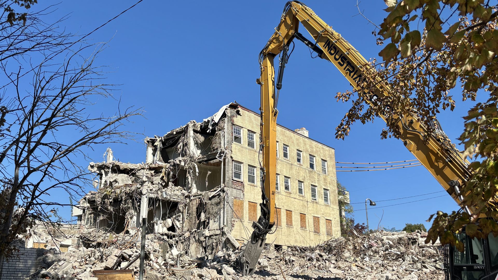 Yellow excavator with a long hydraulic arm tears down a partially collapsed beige brick building under a clear blue sky. Rubble covers the ground; bare trees frame the left, leafy branches the right.