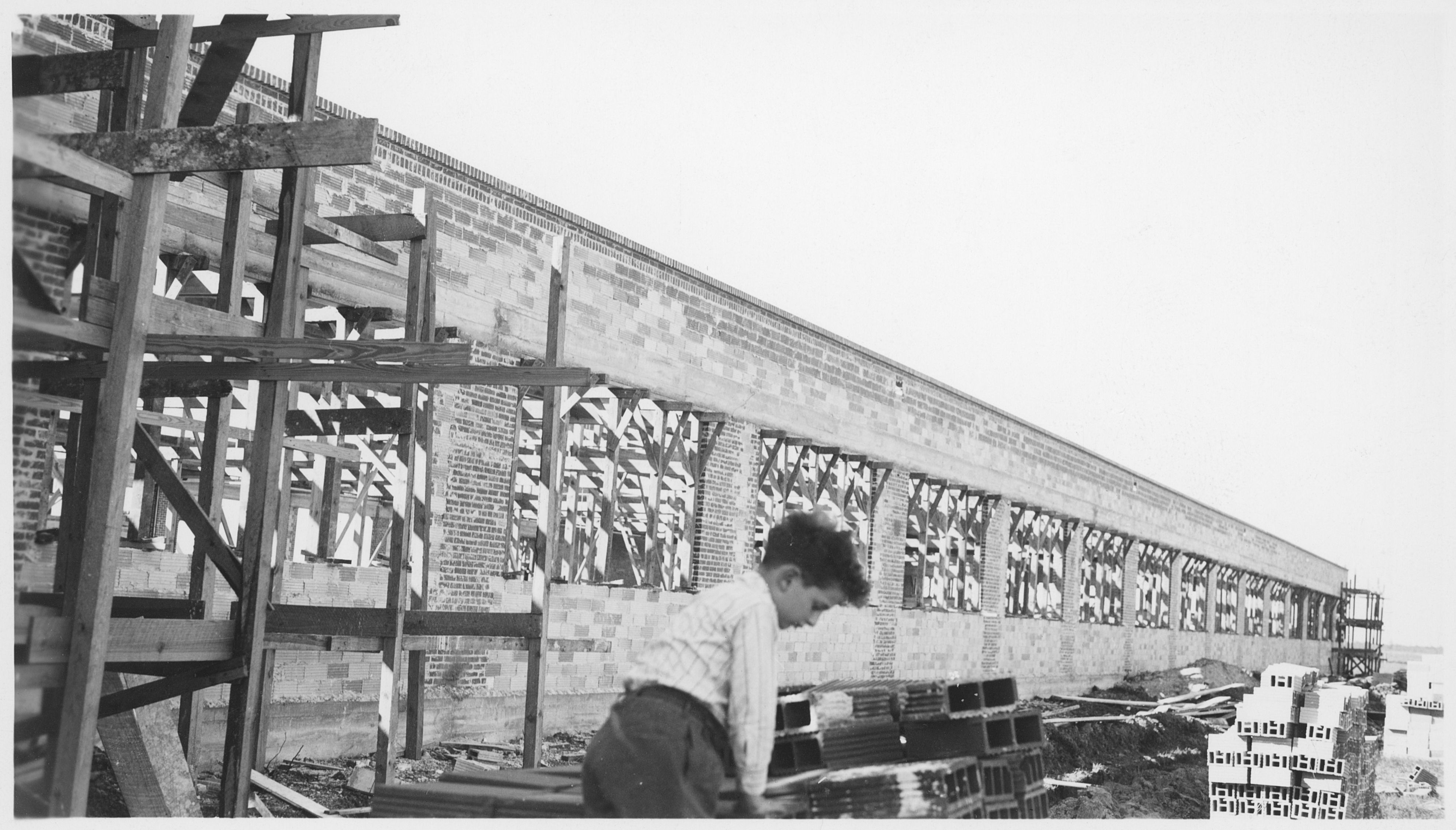 Black-and-white construction site with a long brick wall supported by wooden scaffolding; stacked bricks and a man in a plaid shirt foreground.