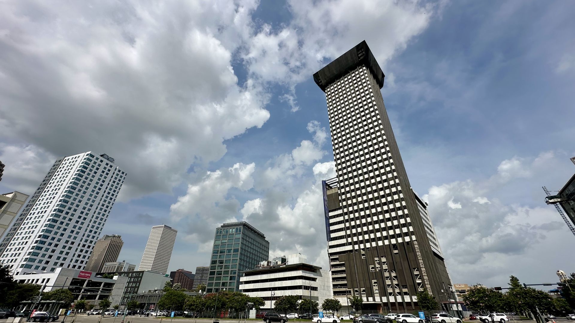 Cityscape with tall modern buildings under a partly cloudy sky, featuring a prominent dark skyscraper with a broad top and several white and glass buildings around it.