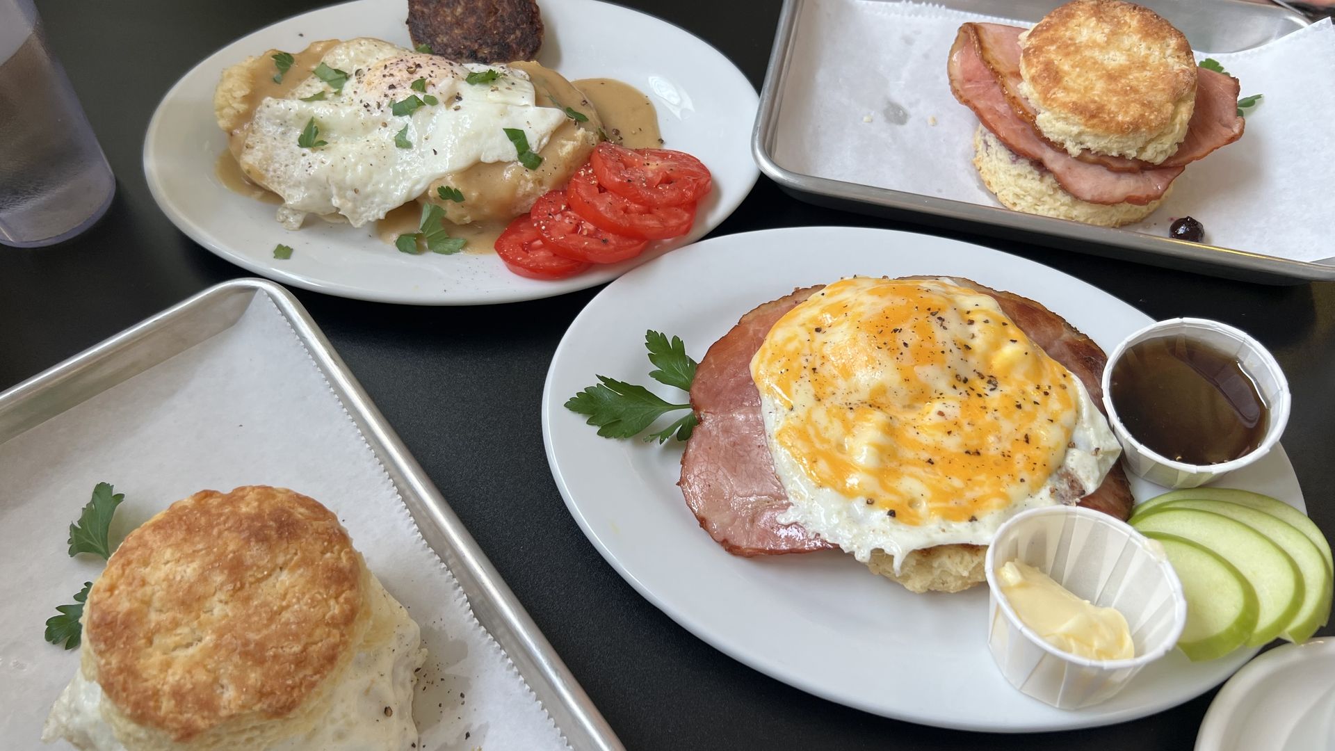 Assorted breakfast on white plates: eggs Benedict with hollandaise and tomato slices; ham-and-egg open-face with cheese; ham biscuit sandwich; apple slices and butter on the side; biscuit on a tray.