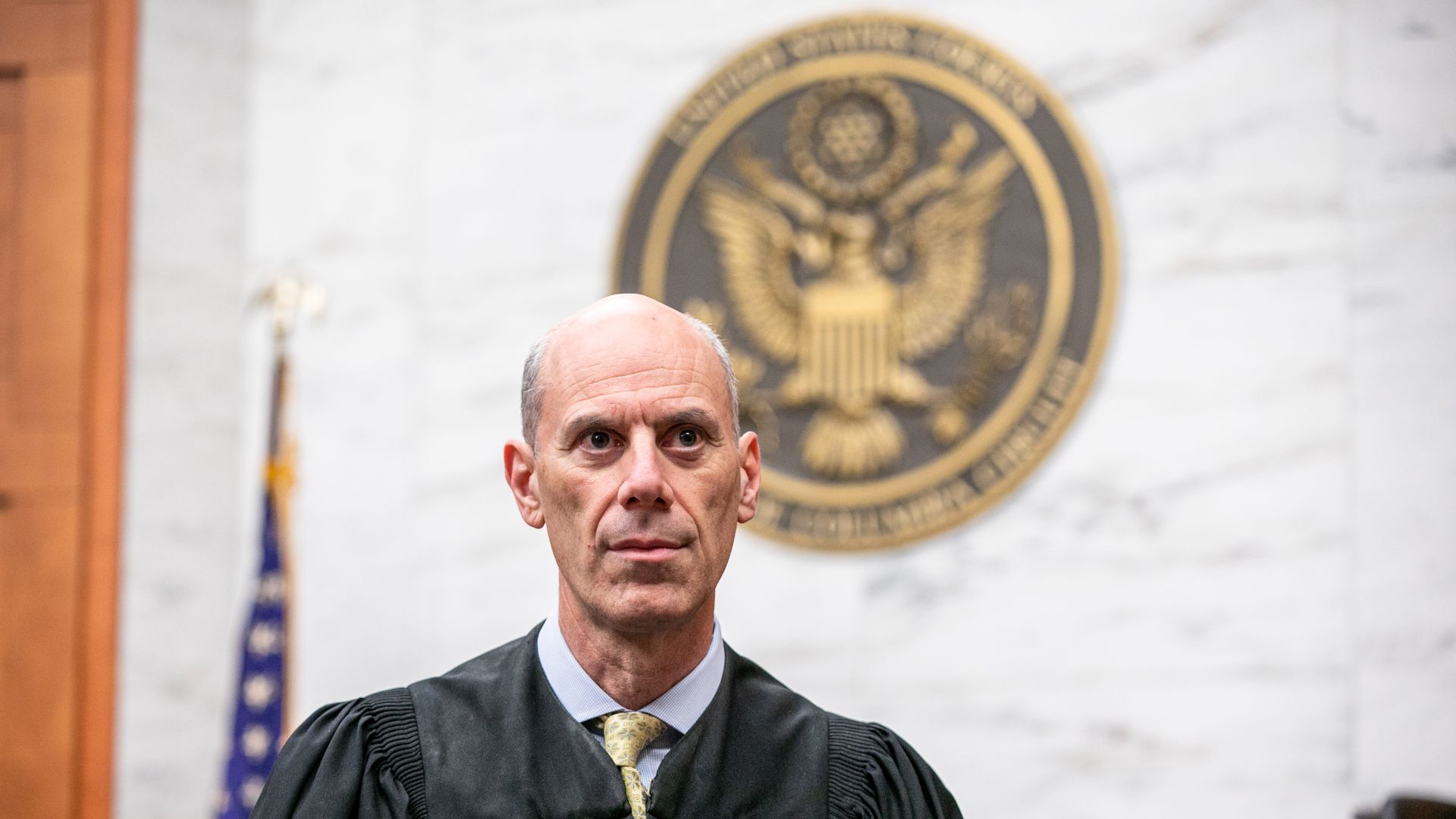 A man in a black judge's robe and yellow patterned tie stands in front of a U.S. government seal and an American flag in a courtroom with marble walls.