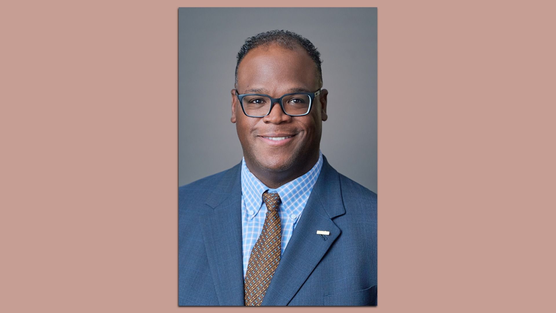 Portrait of a smiling Black man wearing a blue suit, light blue checkered shirt, brown patterned tie, and dark glasses, against a gray studio backdrop.