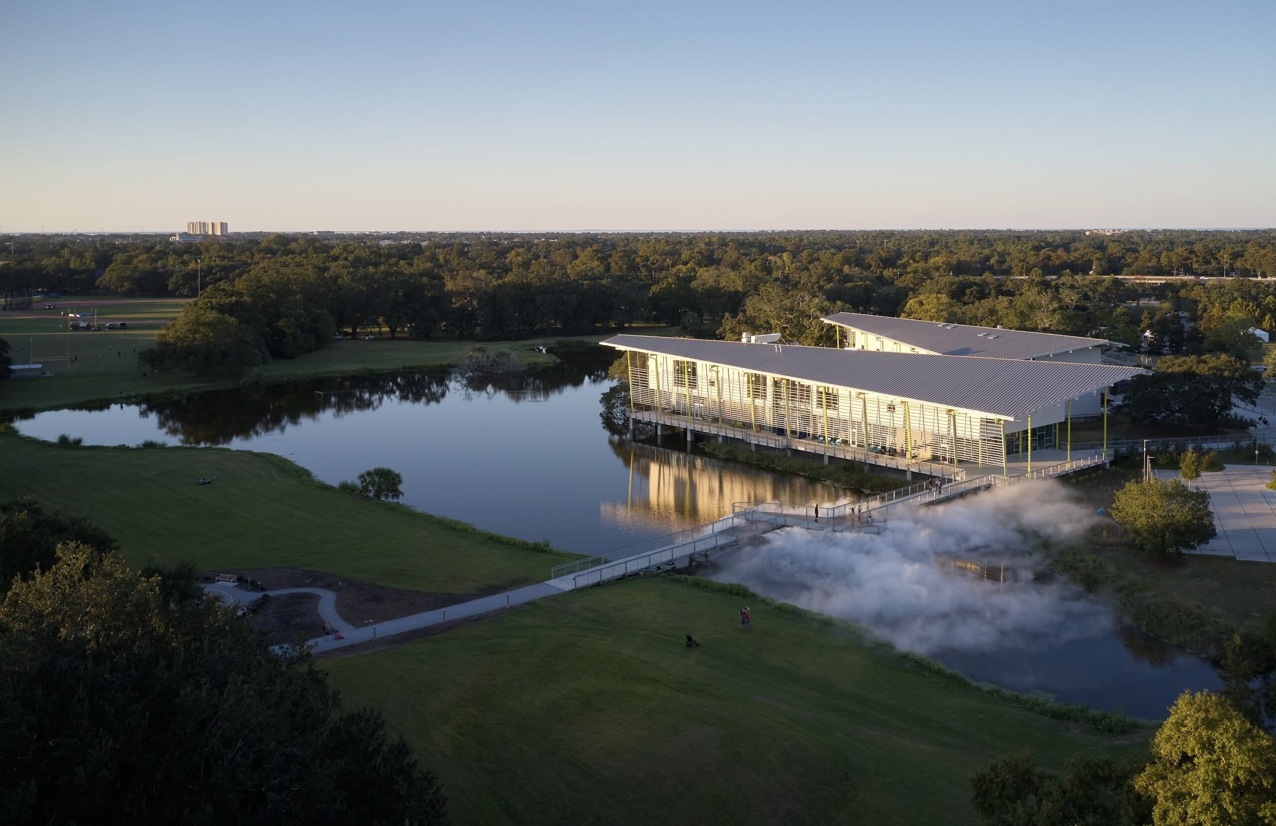 Aerial view of a park beside a calm lake. A long white building with a slanted roof extends over the water while green trees fill the horizon under a clear blue sky. Steam rises near the foreground.