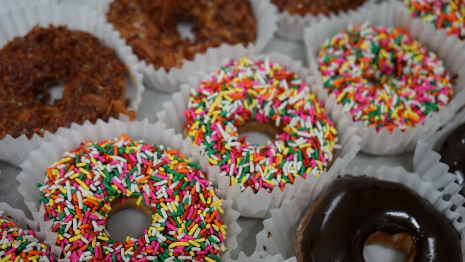 Rows of doughnuts: Coconut on the left, rainbow sprinkles in the middle and chocolate glazed on the right.