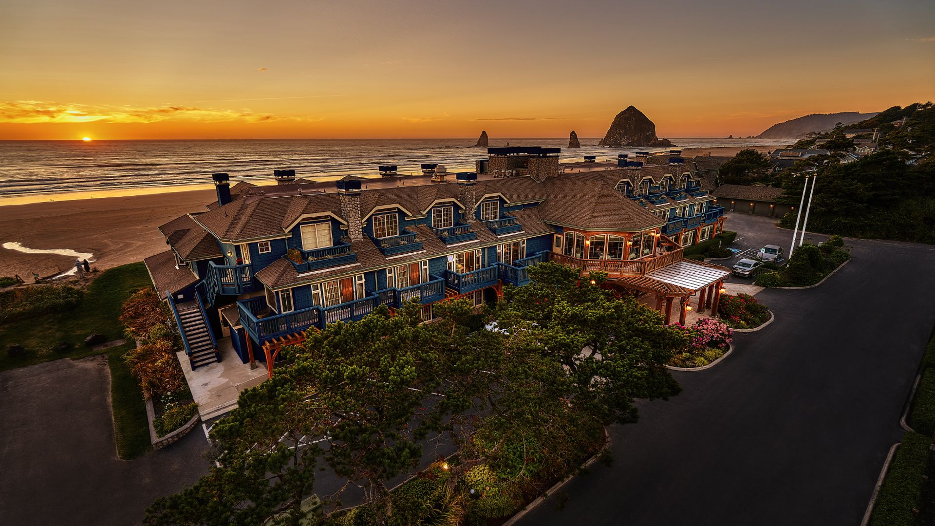 Blue and brown beachfront hotel at sunset with ocean waves, sandy beach, large rock formations, and a few cars in the parking area surrounded by trees and flowers.