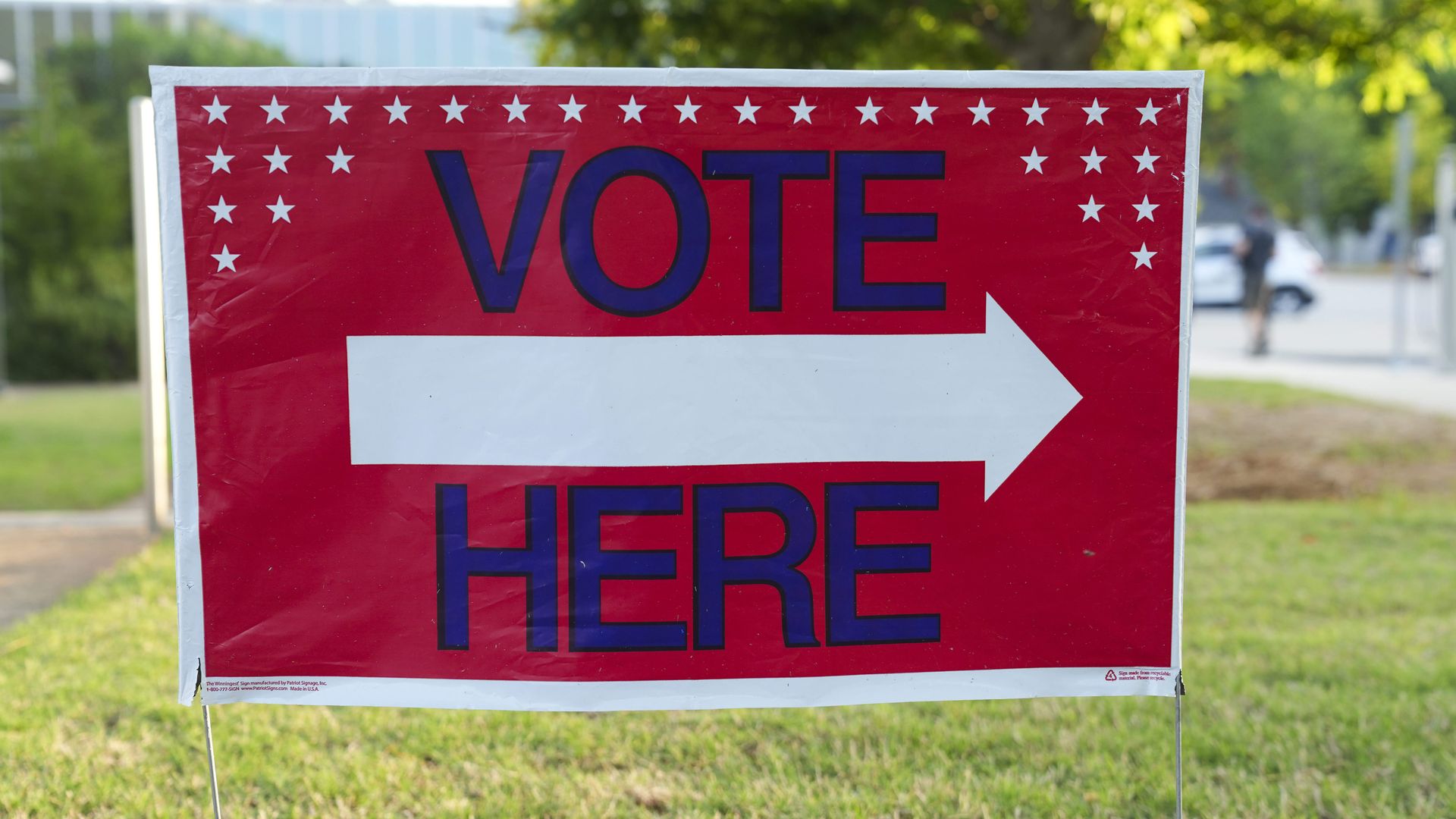 A red sign with dark blue lettering that reads "Vote Here," with small white stars dotted around the upper half of the sign. A white arrow pointing right is position between the words "Vote" and "Here."