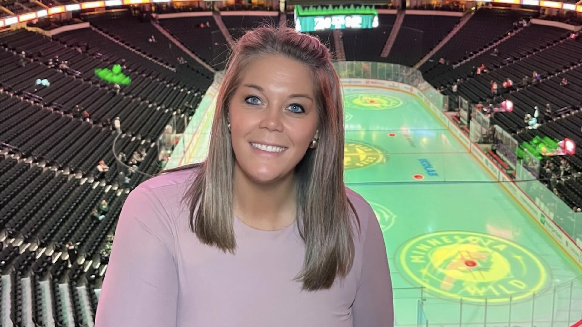 Smiling woman with light brown hair in a pale pink long-sleeve top stands in a large hockey arena, with green-lit stands and a rink displaying a circular logo behind her.