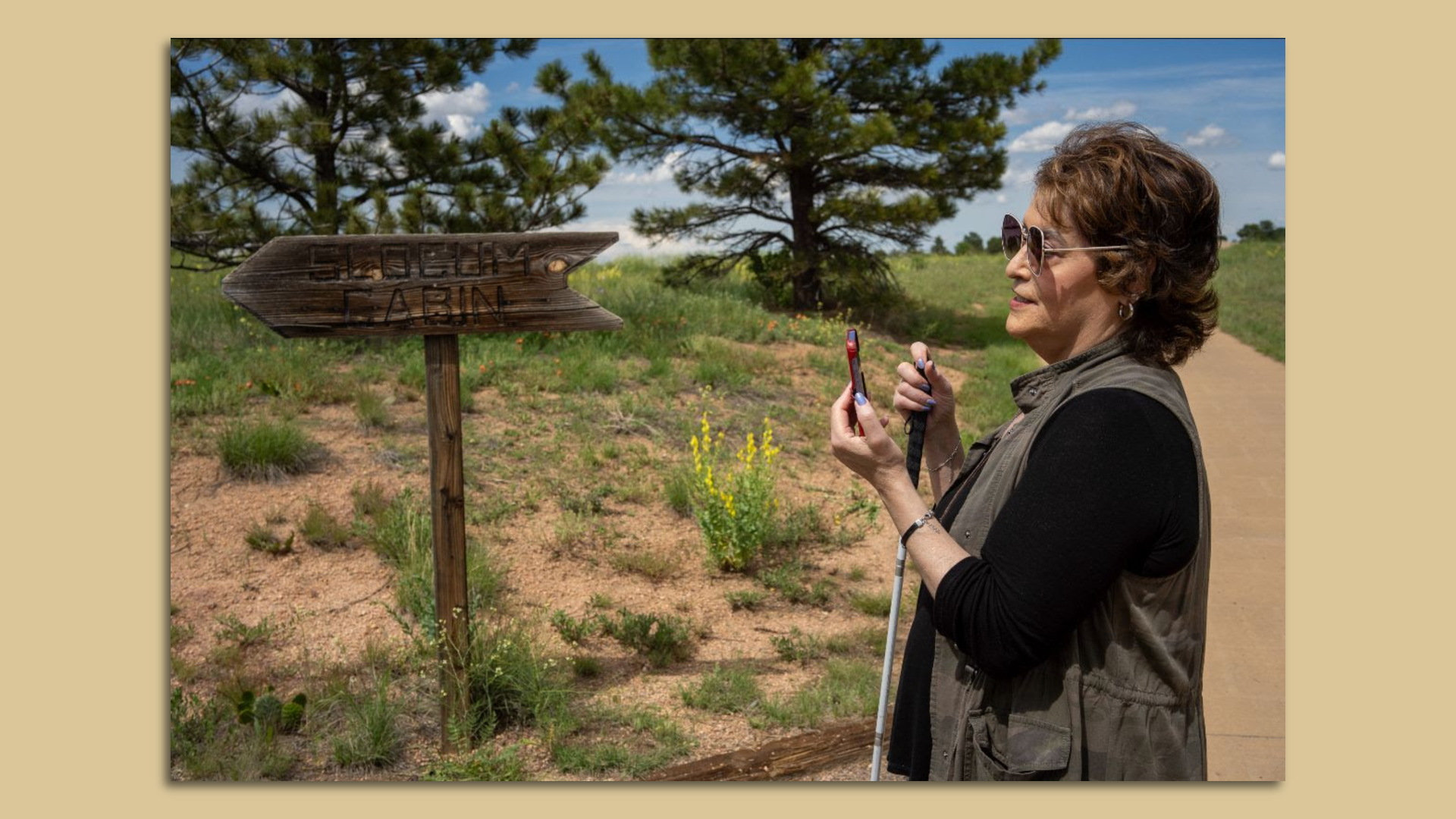 A woman in sunglasses with a mobility cane stands outside and holds her phone up to a state park sign.  