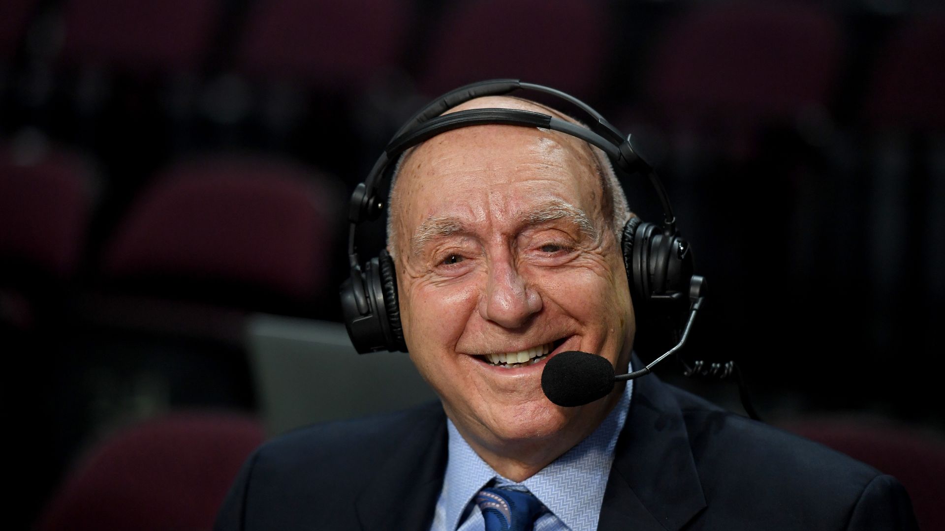 Dick Vitale sits with a microphone in front of his face smiling.