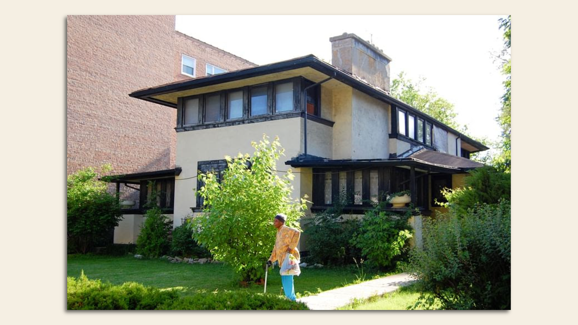 Prairie style house with green grass and a woman in front using a cane.