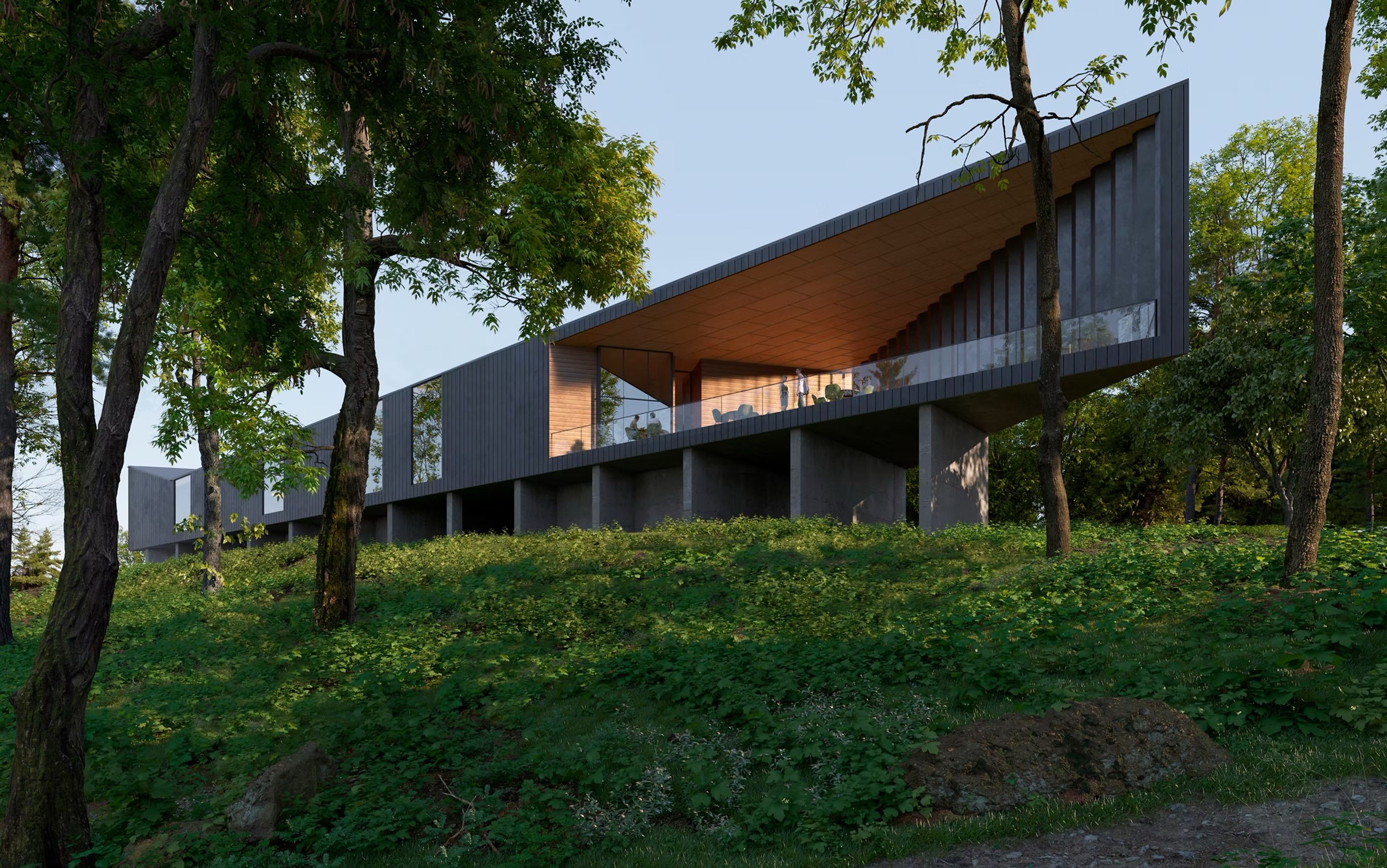 Modern, cantilevered building with a dark gray facade and glass walls sits on stilts in a wooded hillside. A warm-lit interior shows people inside; surrounding trees frame the structure against a blue sky.