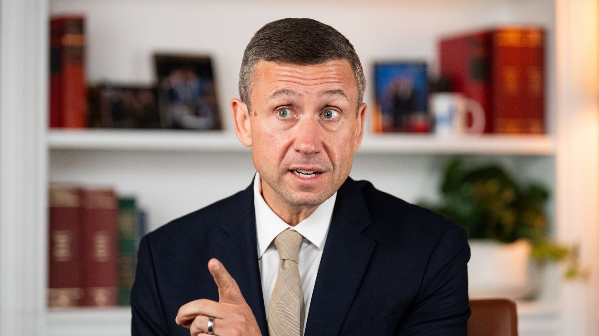 A man in a navy suit and beige tie sits at a desk, gesturing with his index finger. He speaks in a bright home office with a white bookshelf, red books, framed photos, and a plant nearby.