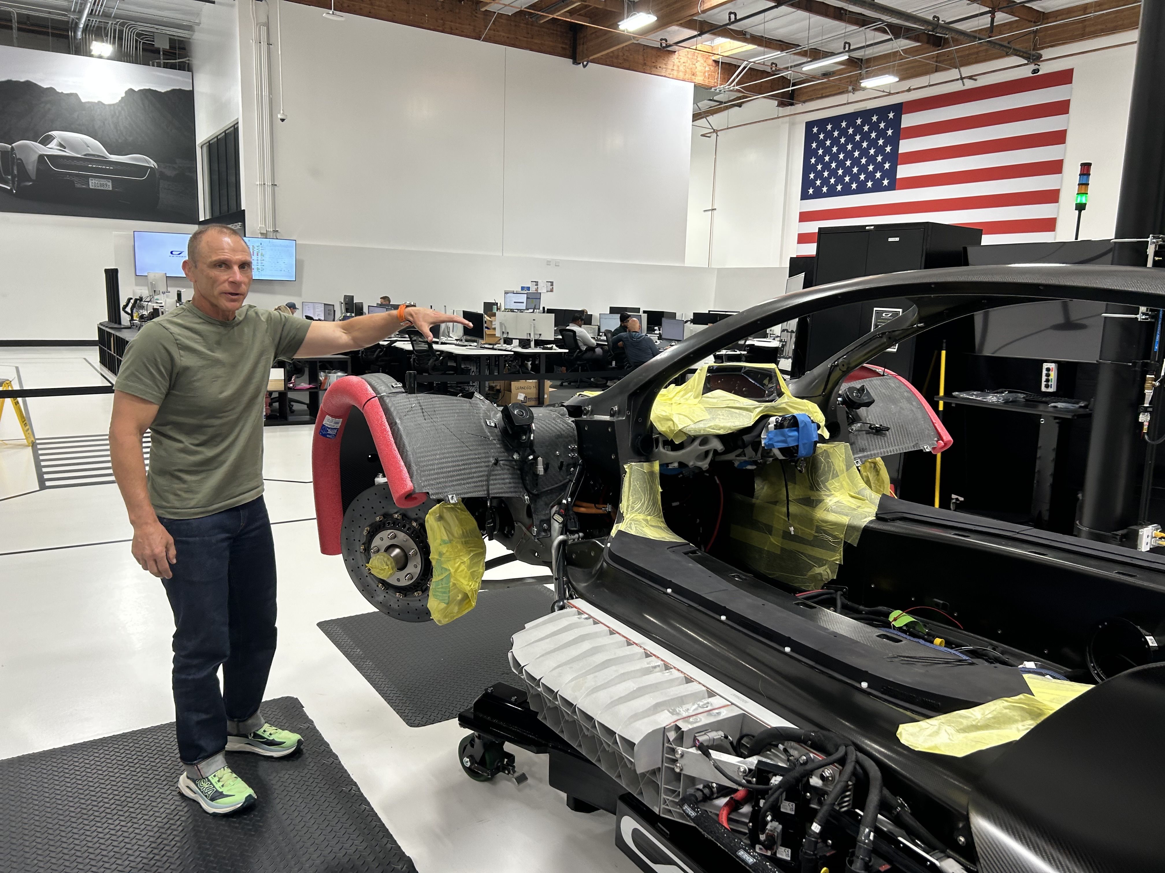 Man in a green T-shirt points at the exposed cockpit of a partially assembled black sports car in a bright workshop; yellow tape covers parts, and an American flag hangs on the wall.