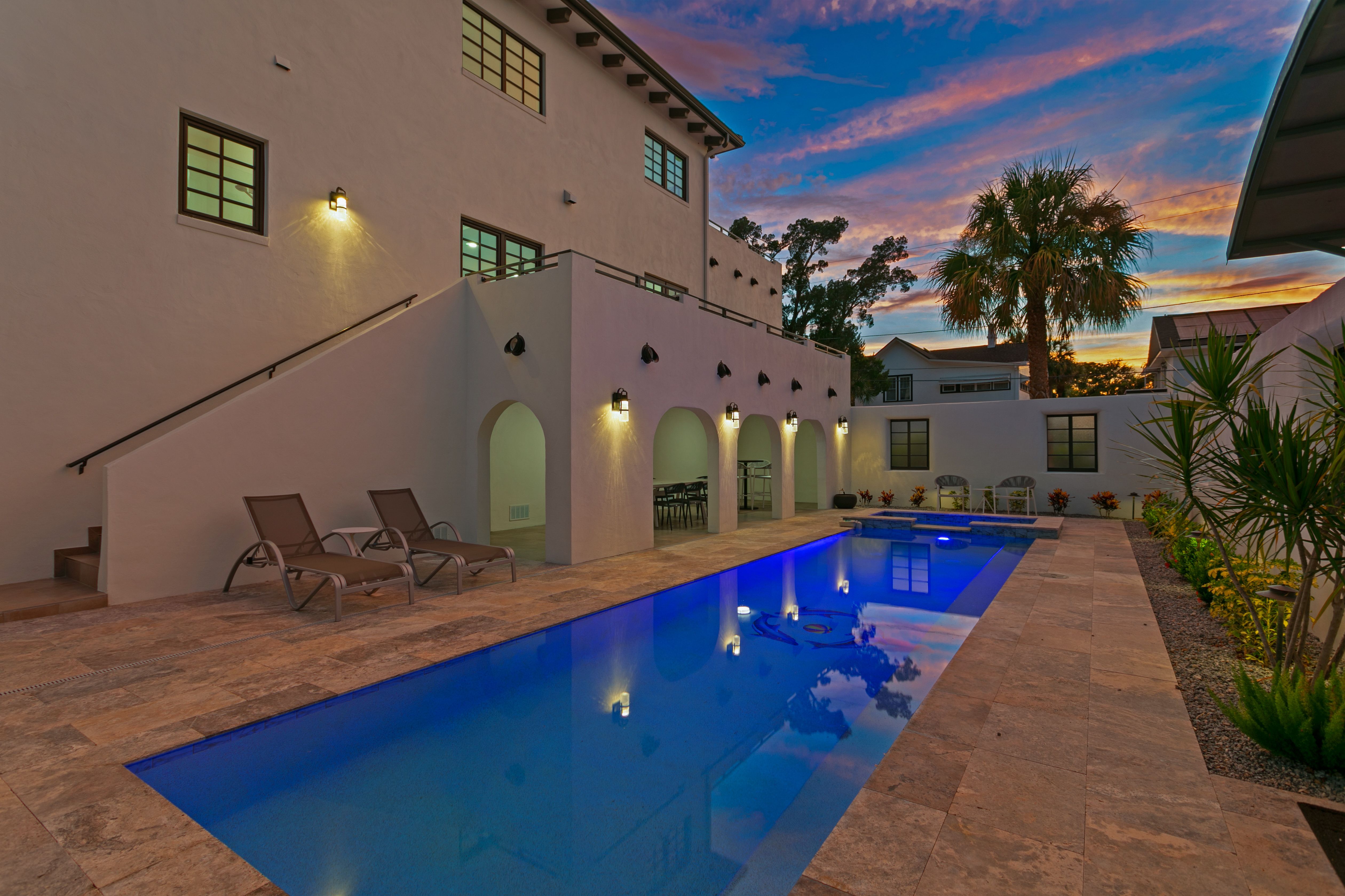 A deep blue lap pool pictured in the evening with a blue-and-pink sunset in the sky above. The pool is surrounded by sand-colored tile and two chaise lounges.