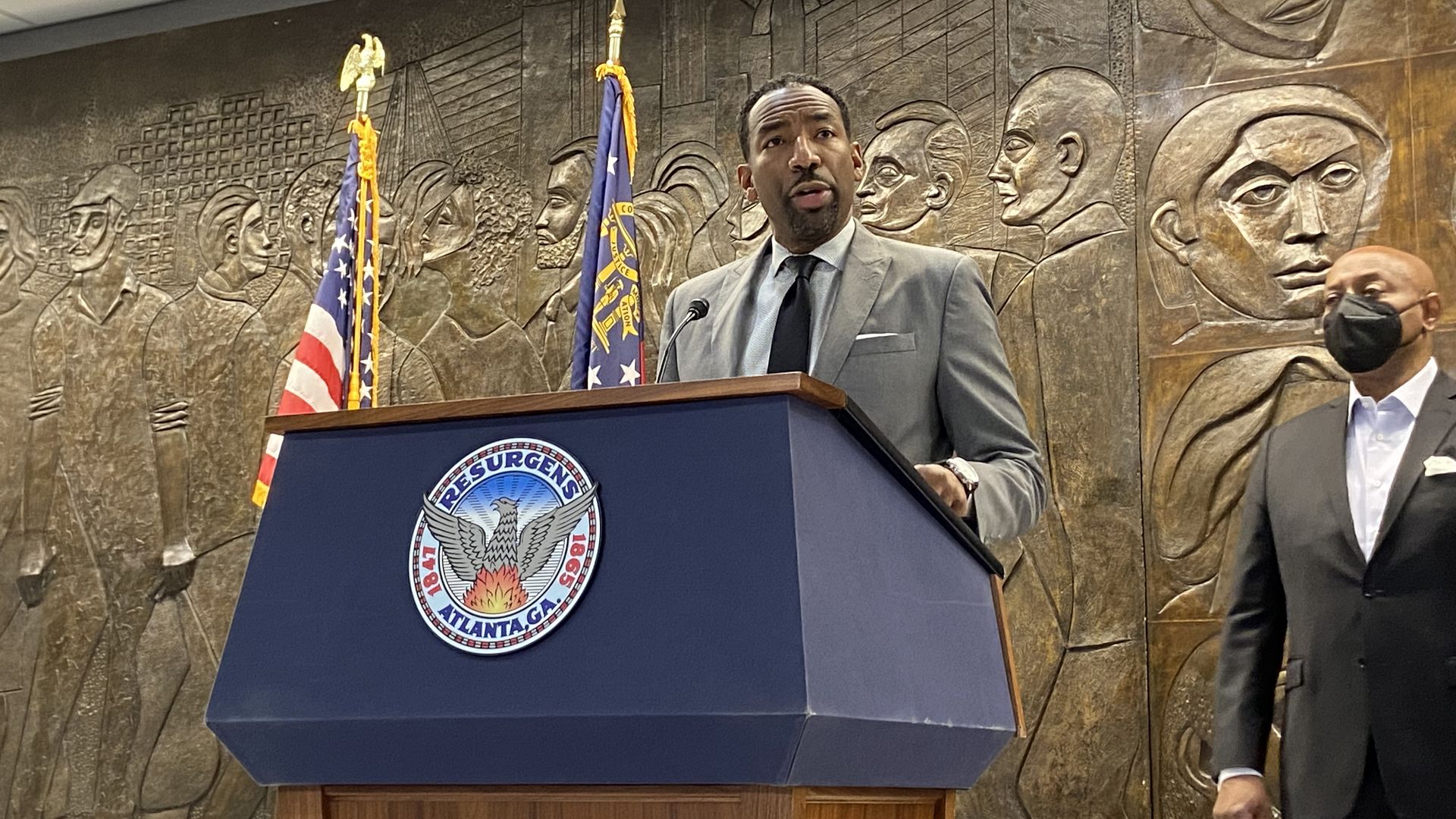 Mayor Andre Dickens, a tall man wearing a gray suit, stands at a lectern with the Atlanta city seal and in front of a bas relief wall sculpture of people's faces