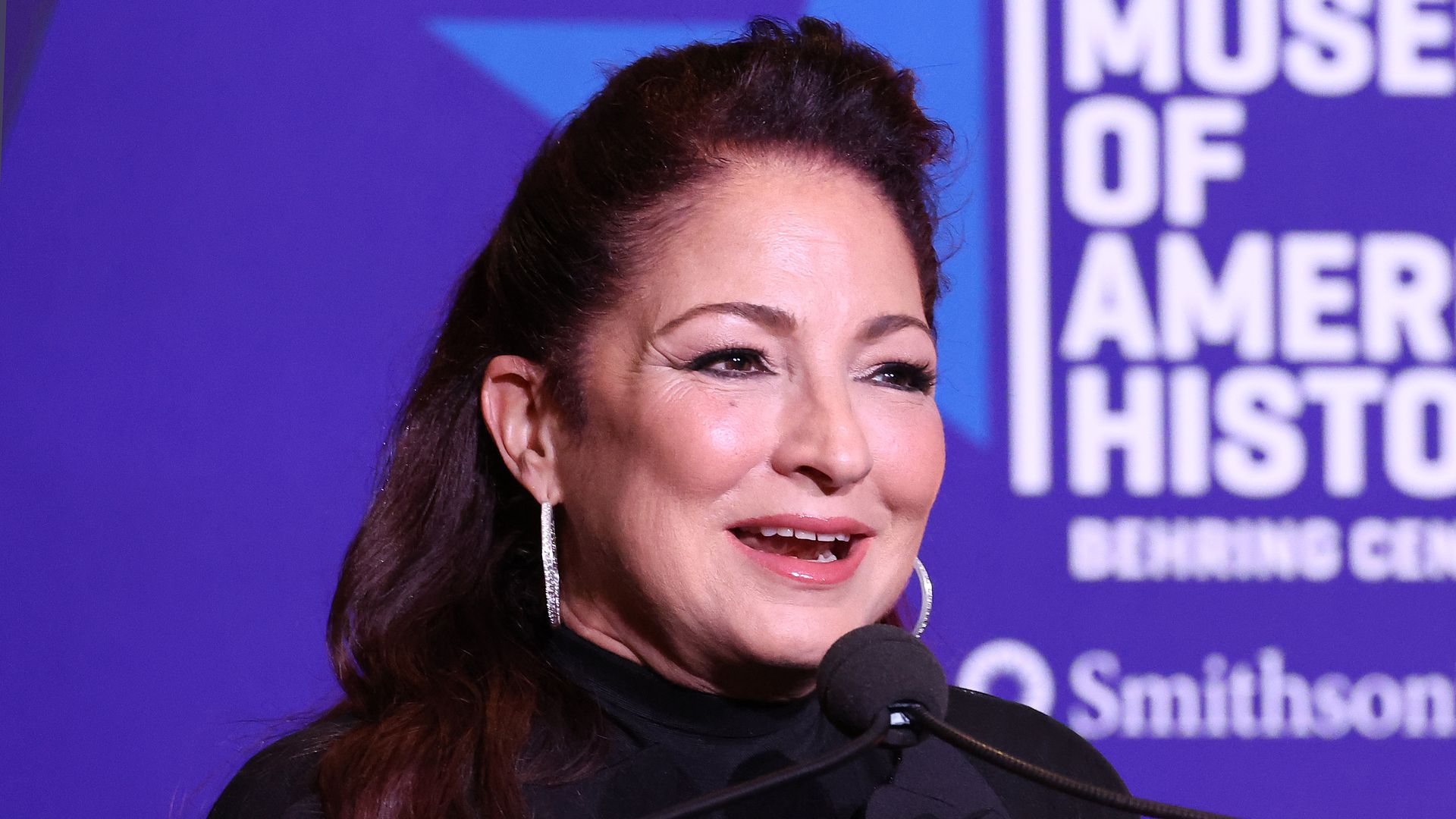 Gloria Estefan smiles while in front of a a purple background 