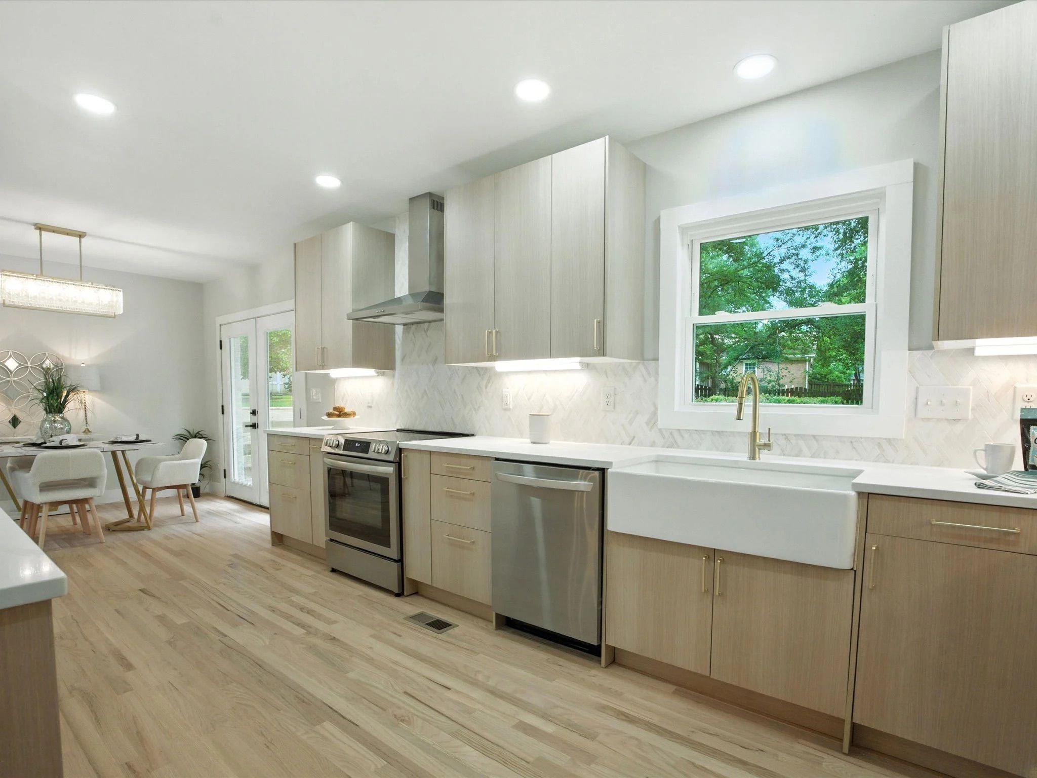 Bright modern kitchen with light wood cabinets, white countertops, stainless steel appliances, farmhouse sink, brass faucet, hardwood floors, and a dining area with white chairs and a pendant light.