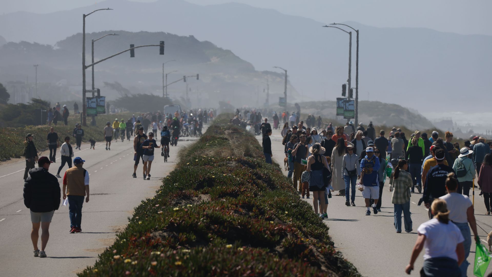 Busy coastal promenade filled with walkers and cyclists, a flowering median in the foreground, sea on the right, hazy hills in the distance, and streetlights lining the road.