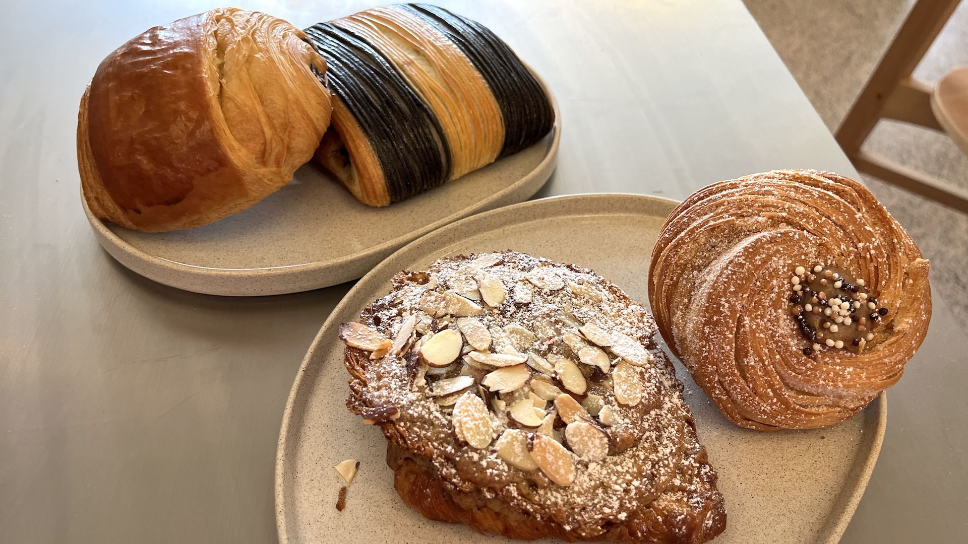 On a light table, a plate holds a long rainbow-striped laminated bread loaf. In the foreground are two sugar-dusted pastries—one almond-topped round, the other a swirled pastry with pearl sugar.