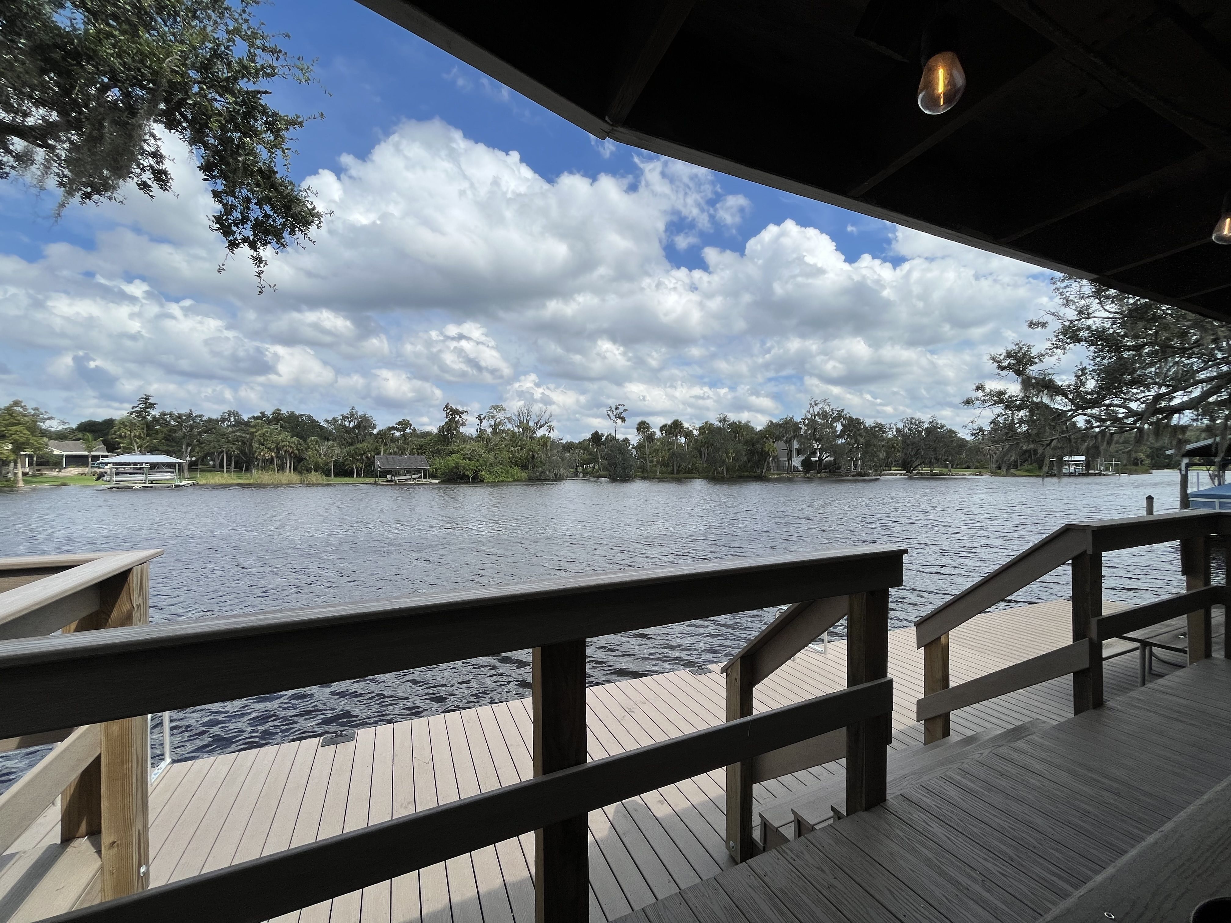 View from a shaded wooden deck over a calm lake, with brown railing, tree-lined shore, boats on docks, and a blue sky with white fluffy clouds.