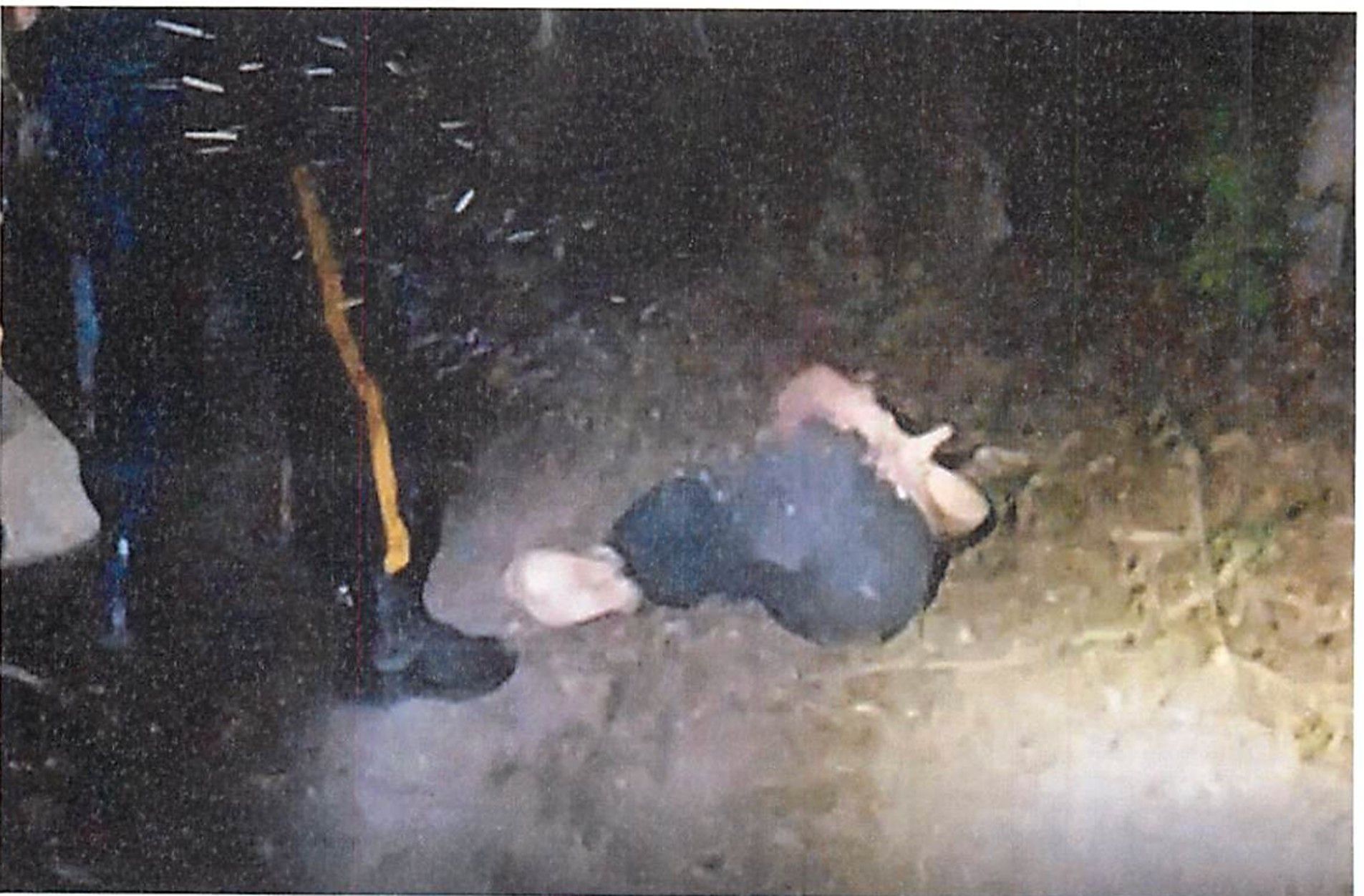Person in dark clothing lying on the ground at night next to someone wearing black pants with a yellow stripe, on a dirt surface with some vegetation.