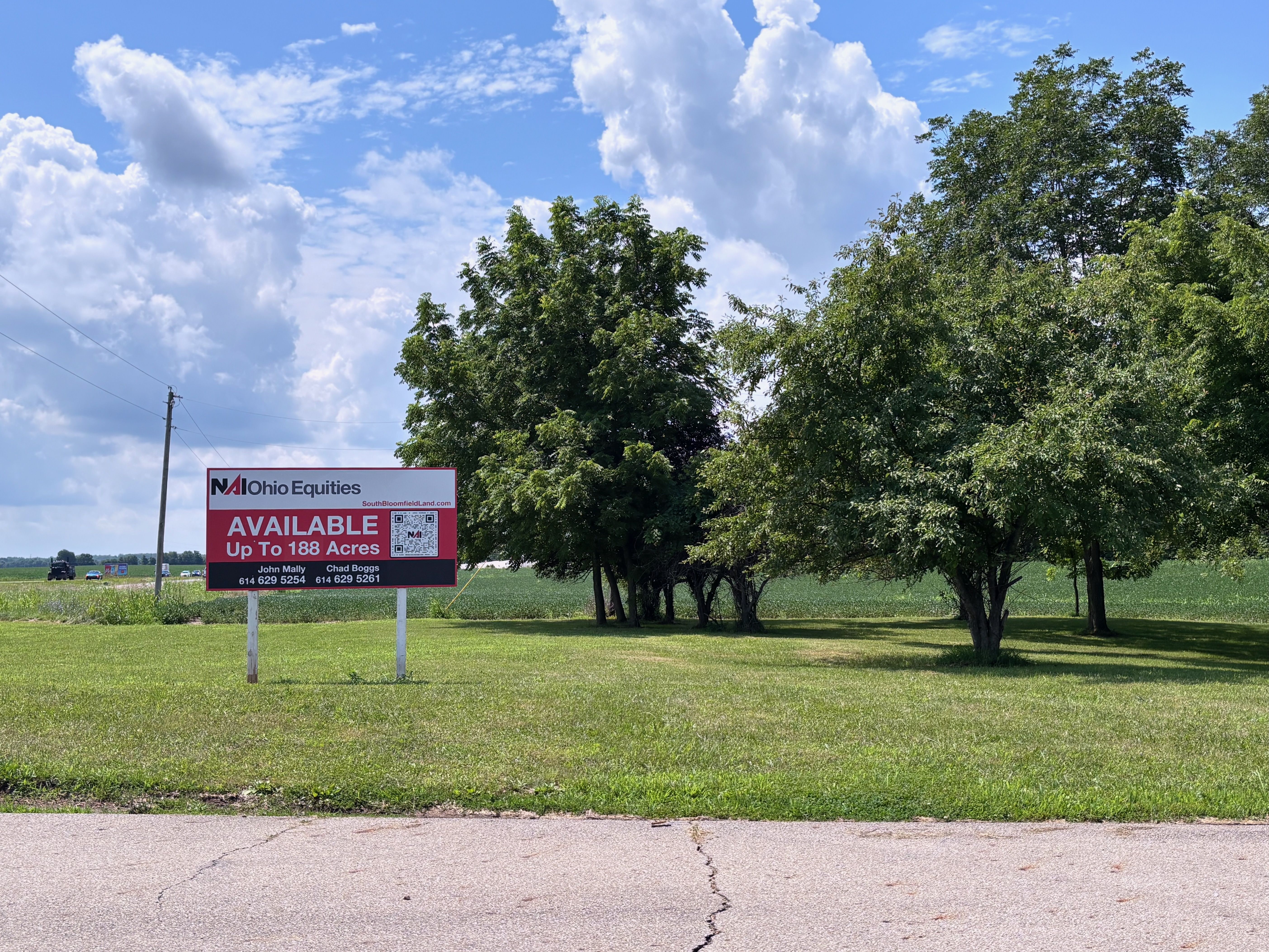 Grassy field with a line of green trees to the right, a bright blue sky with white clouds, and a red-and-white sign in the foreground advertising available land up to 188 acres; a utility pole and distant cars on a road.