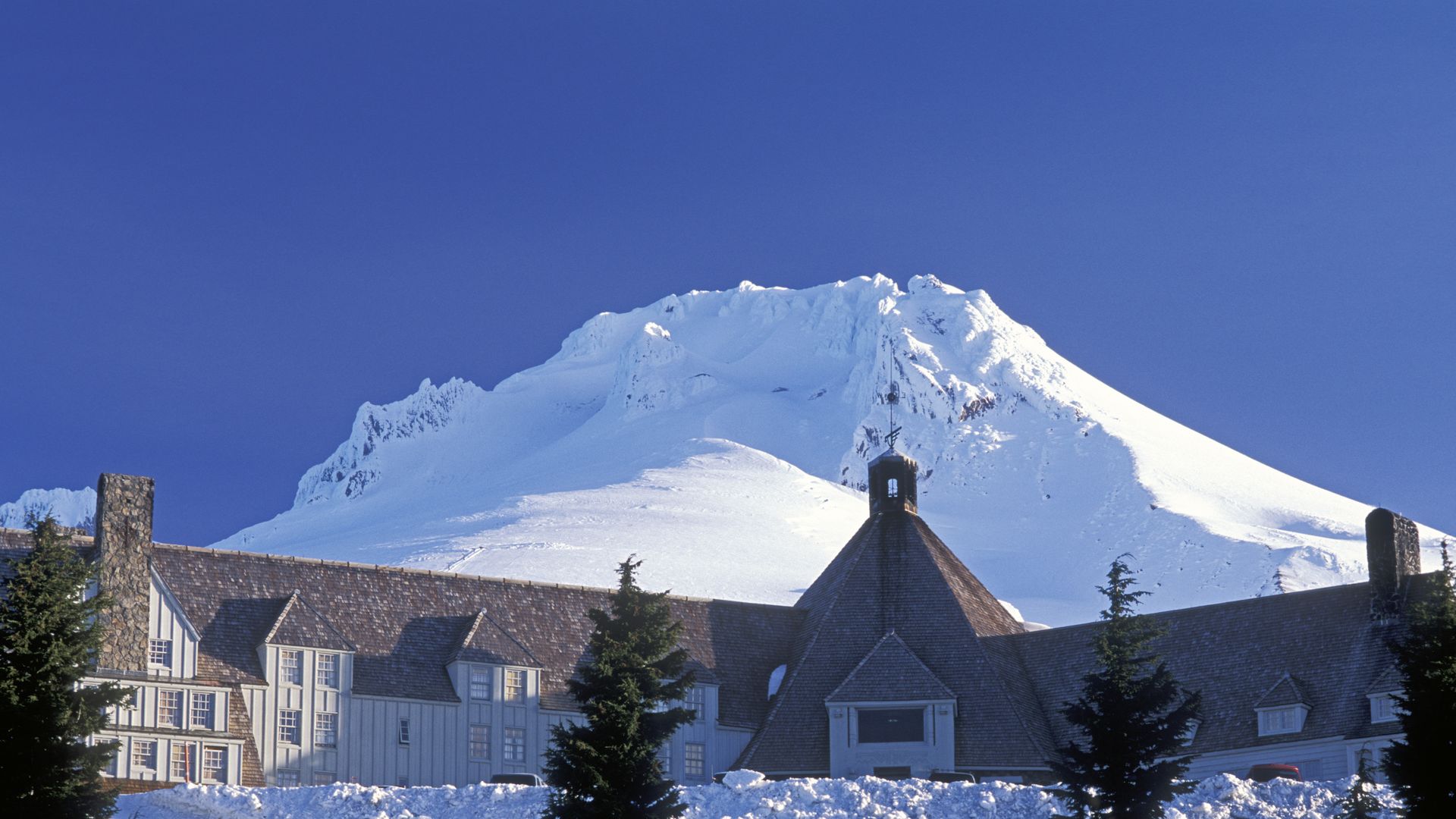 Snow-covered mountain under clear blue sky behind a large lodge-style building with peaked roofs and stone chimneys, surrounded by snow and evergreen trees.