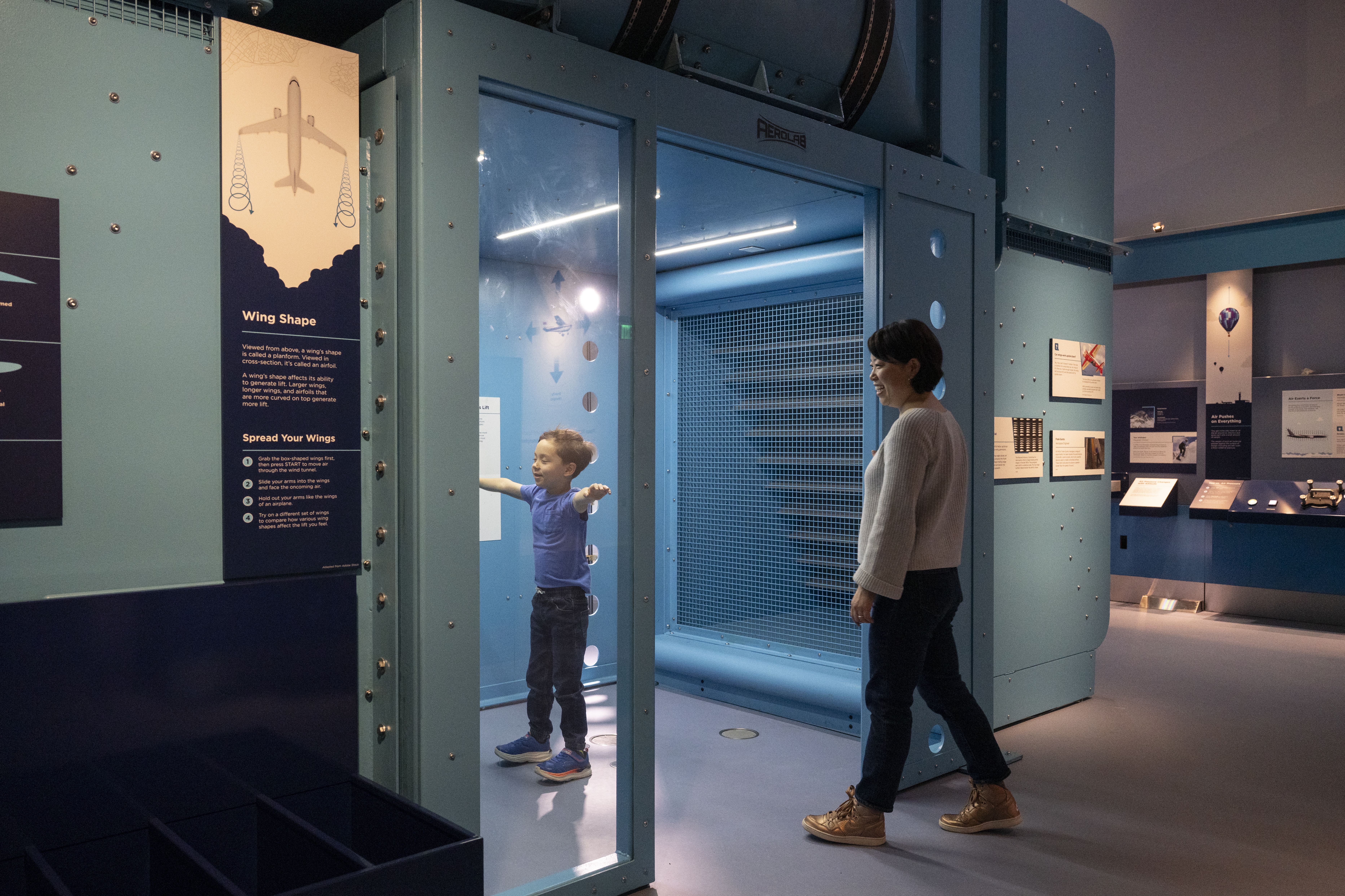 Exhibit with a blue aircraft wing display. A boy in a blue shirt stands inside a glass chamber, arms outstretched, as a woman watches nearby. A poster titled "Wing Shape" is on the left.