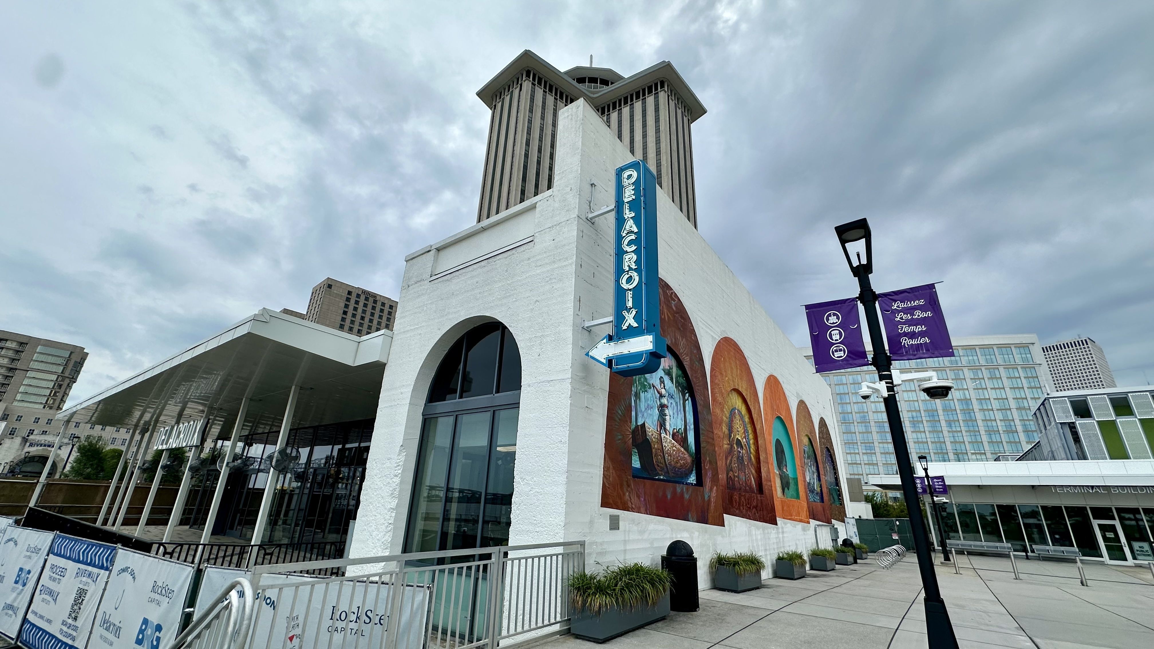 White building with blue "DELACROIX" vertical neon sign and colorful murals under cloudy sky; modern buildings and purple banners saying "Laissez Les Bon Temps Rouler" visible in urban plaza.