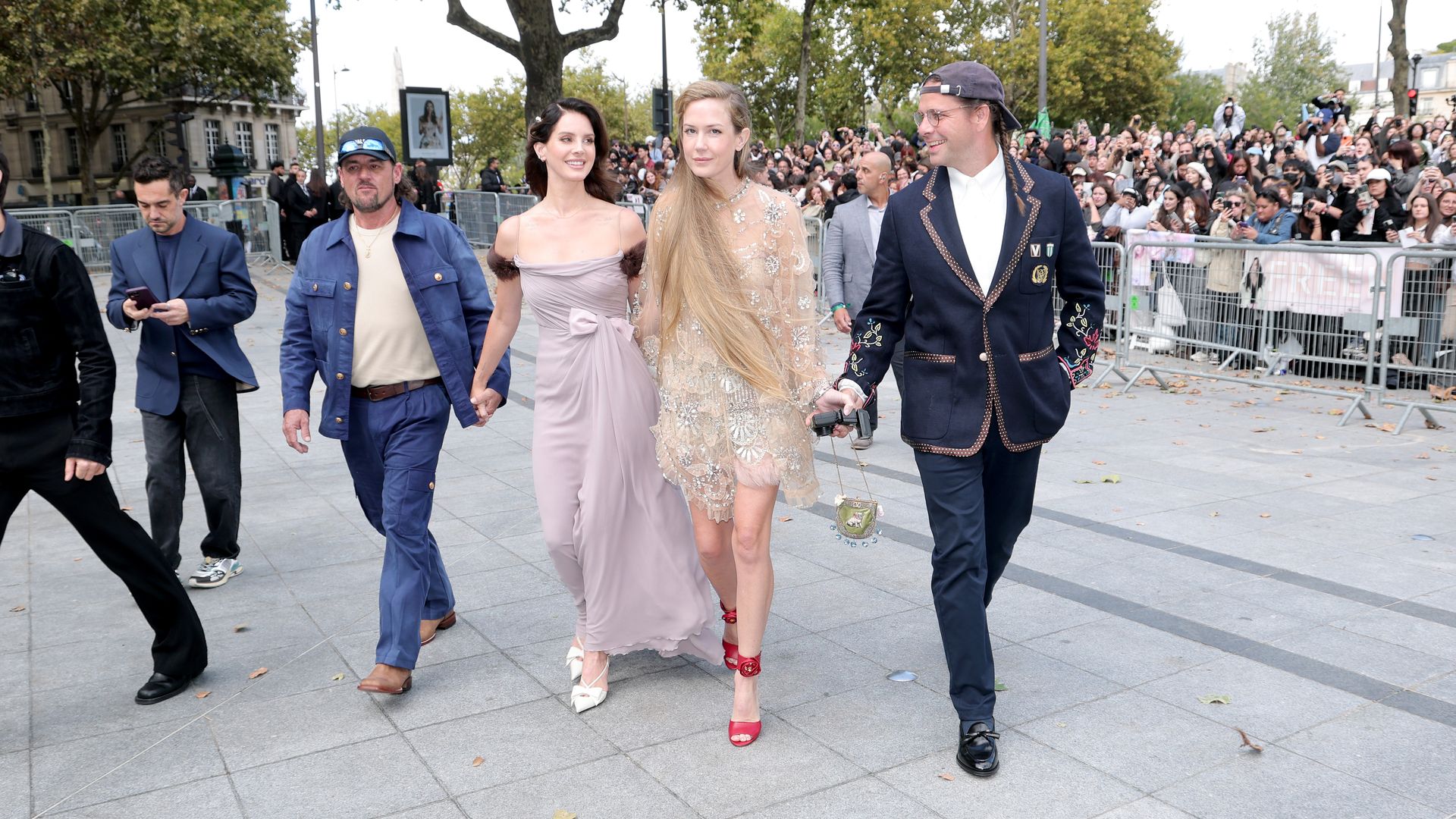 Four people walk, holding hands, across an open plaza in front of barricades, behind which a shoulder-to-shoulder crowd of photographers angles for a closer look.
