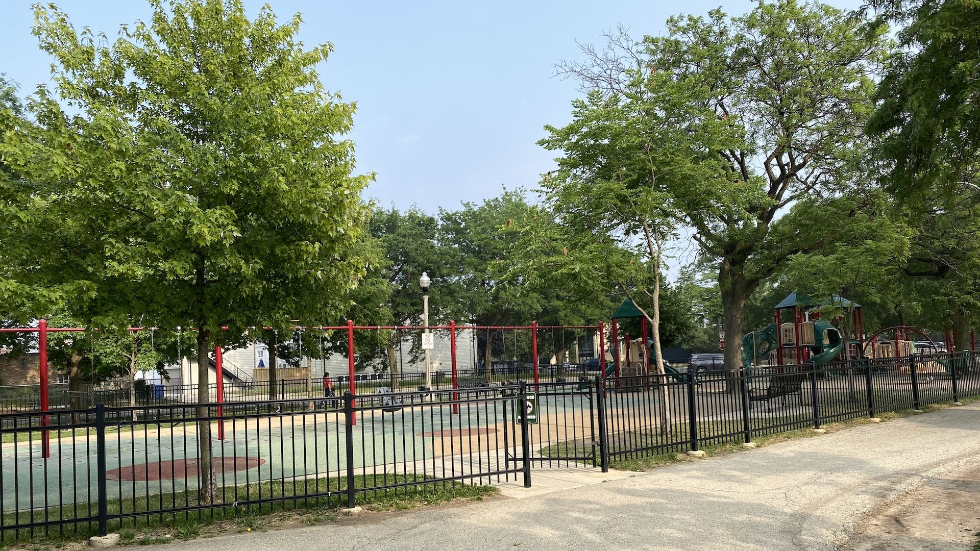 Playground with red swings, green slides, and black fence surrounded by large green trees under a clear blue sky on a sunny day.