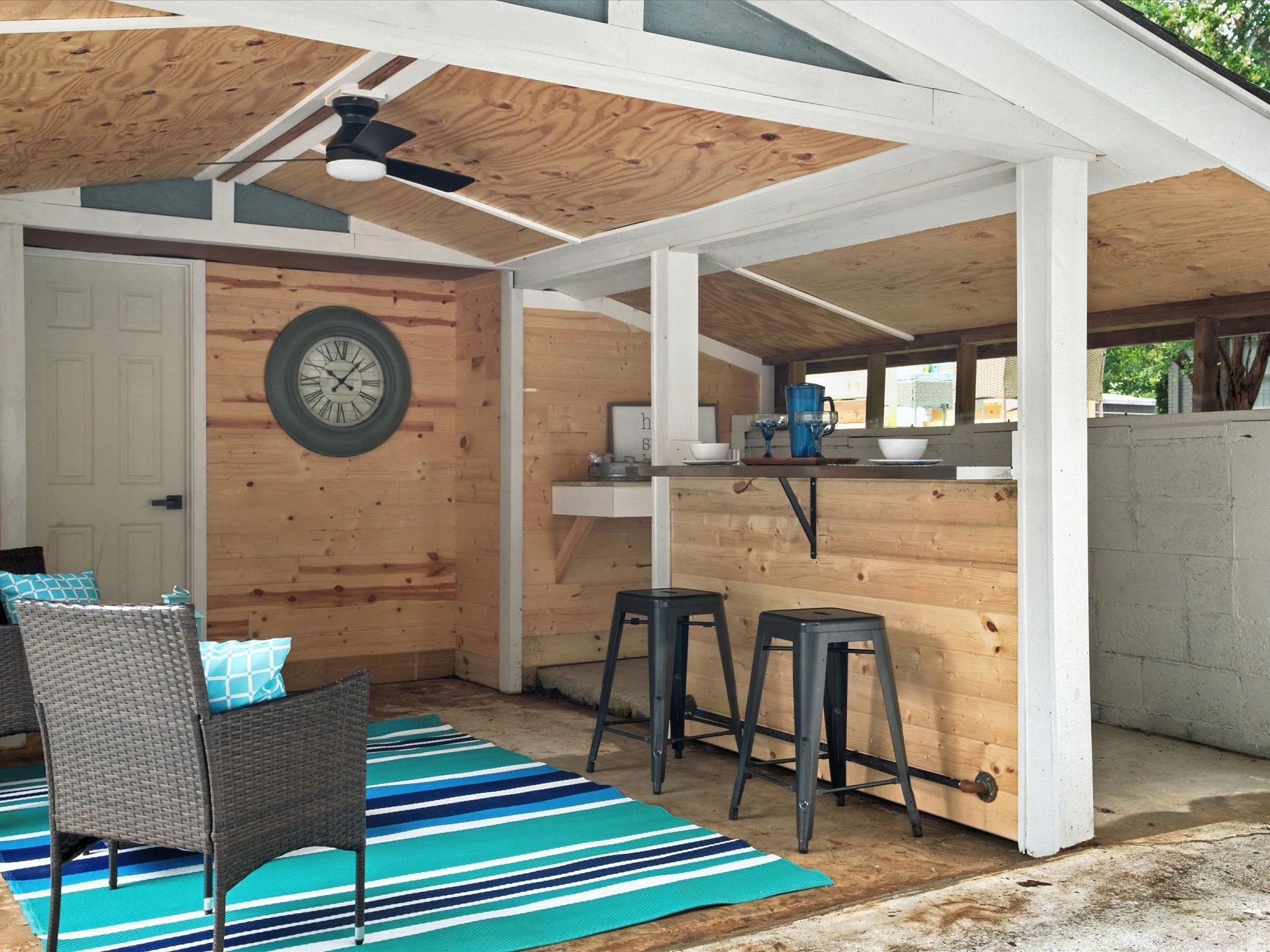Cozy covered patio with wooden walls and ceiling, gray wicker chairs with turquoise pillows, blue-striped rug, two black bar stools at a wood counter, and a round wall clock.
