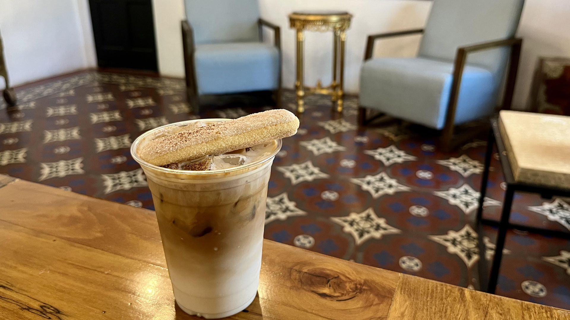 Iced coffee in a plastic cup with a cookie on top sits on a wooden counter. In the background, two light-blue chairs flank a gold ornate table and framed artwork.
