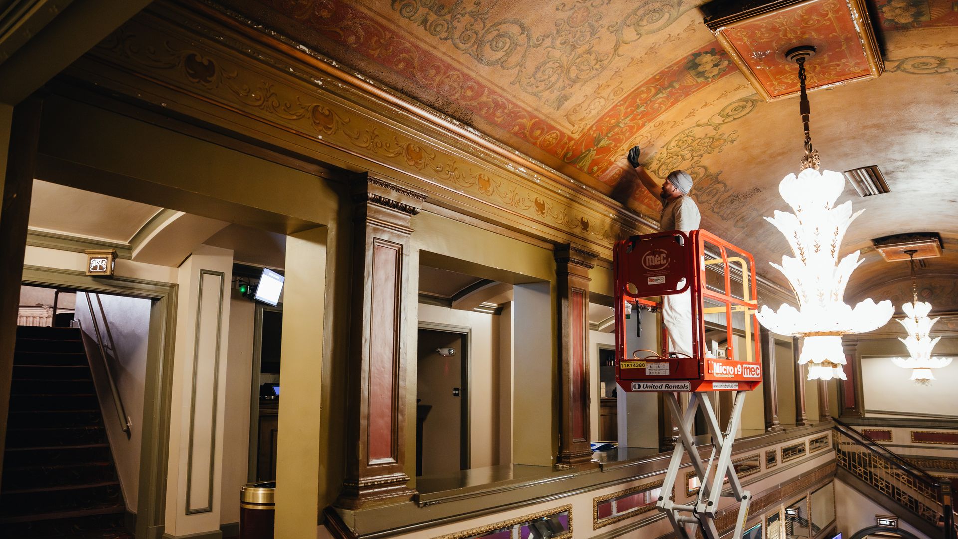 Worker in a gray hard hat on a red lift works on an ornate gilded ceiling in a historic interior, with decorative murals, a bright chandelier, and a grand staircase nearby.