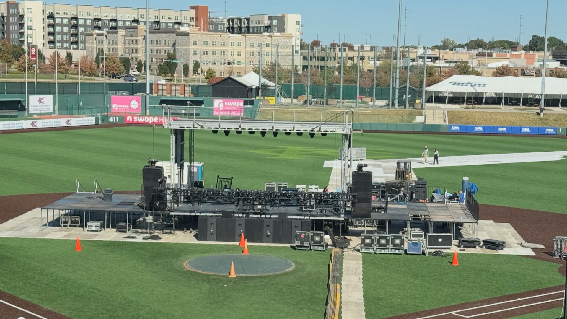 Outdoor stage setup in the infield at Legends Field in KCK with speakers, lighting rigs, and audio equipment surrounded by orange cones and buildings in the background under clear sky.
