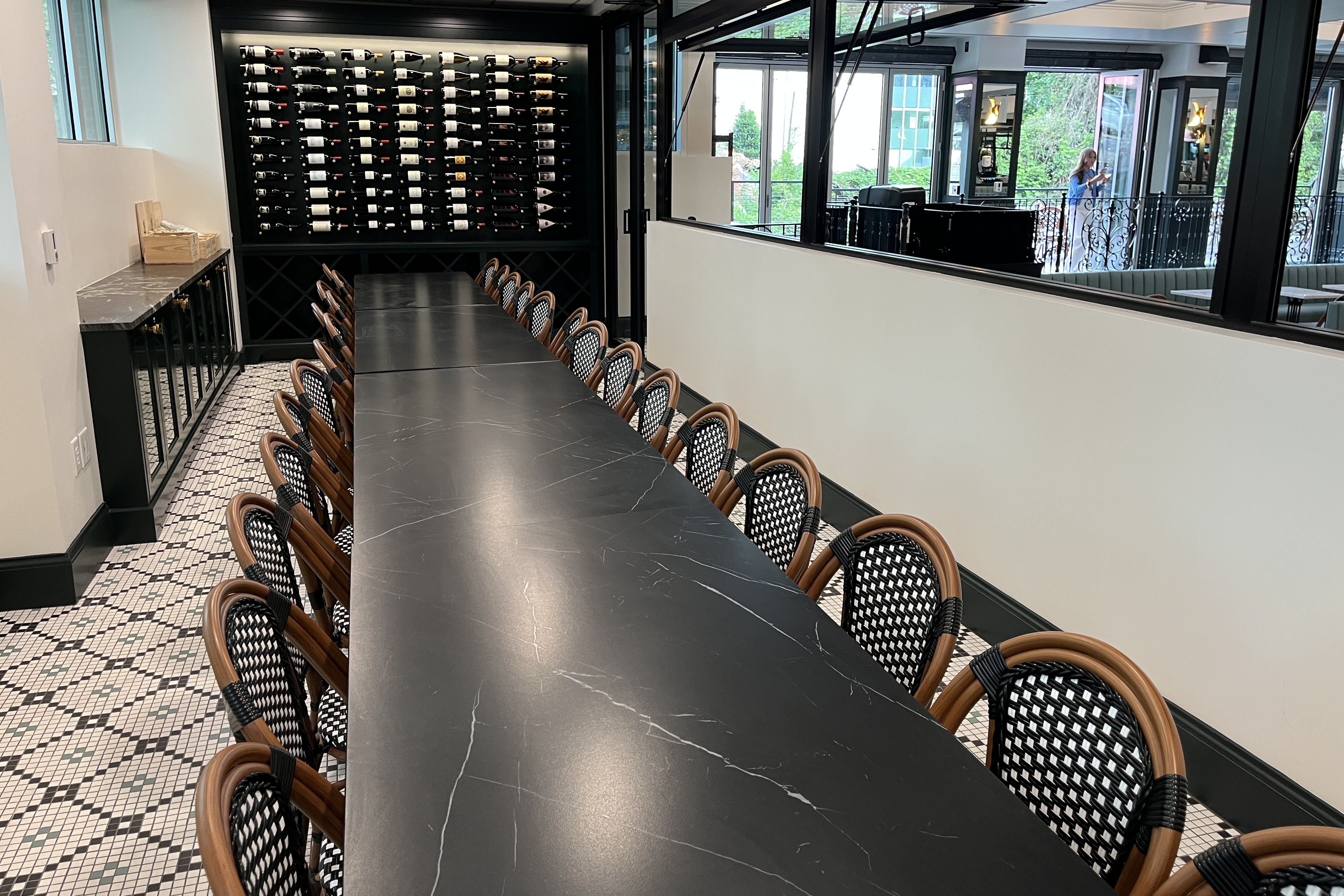 Interior wine-tasting room with a long black marble table and wicker chairs, wall of wine bottles, white walls, and a mosaic tile floor; glass doors overlook greenery outside.