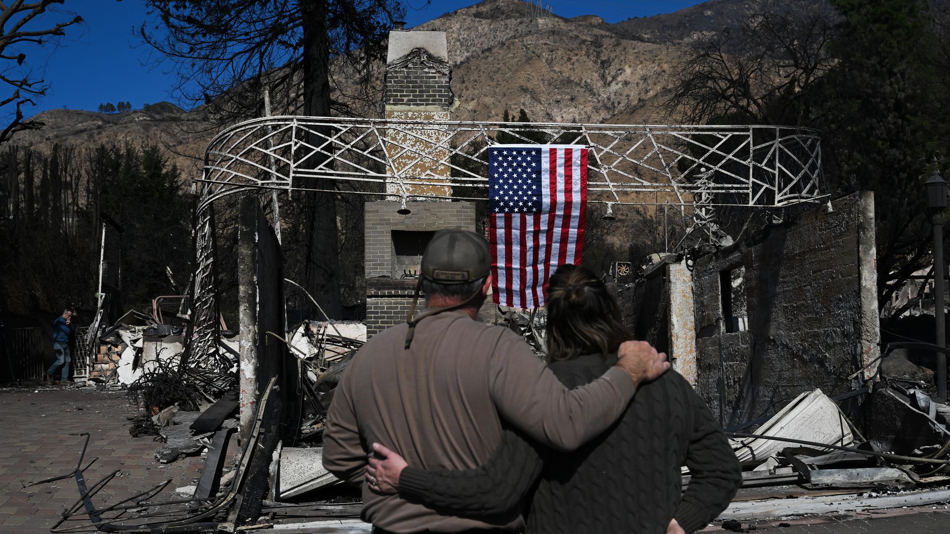 Brian McMahon (left) and Emily Philips (right) look through their remembrances of burned Altadena home as they hung a U.S. flag on debris after massive wildfire in Los Angeles, California, United States on January 13, 2025. (Photo by Tayfun Coskun/Anadolu via Getty Images)