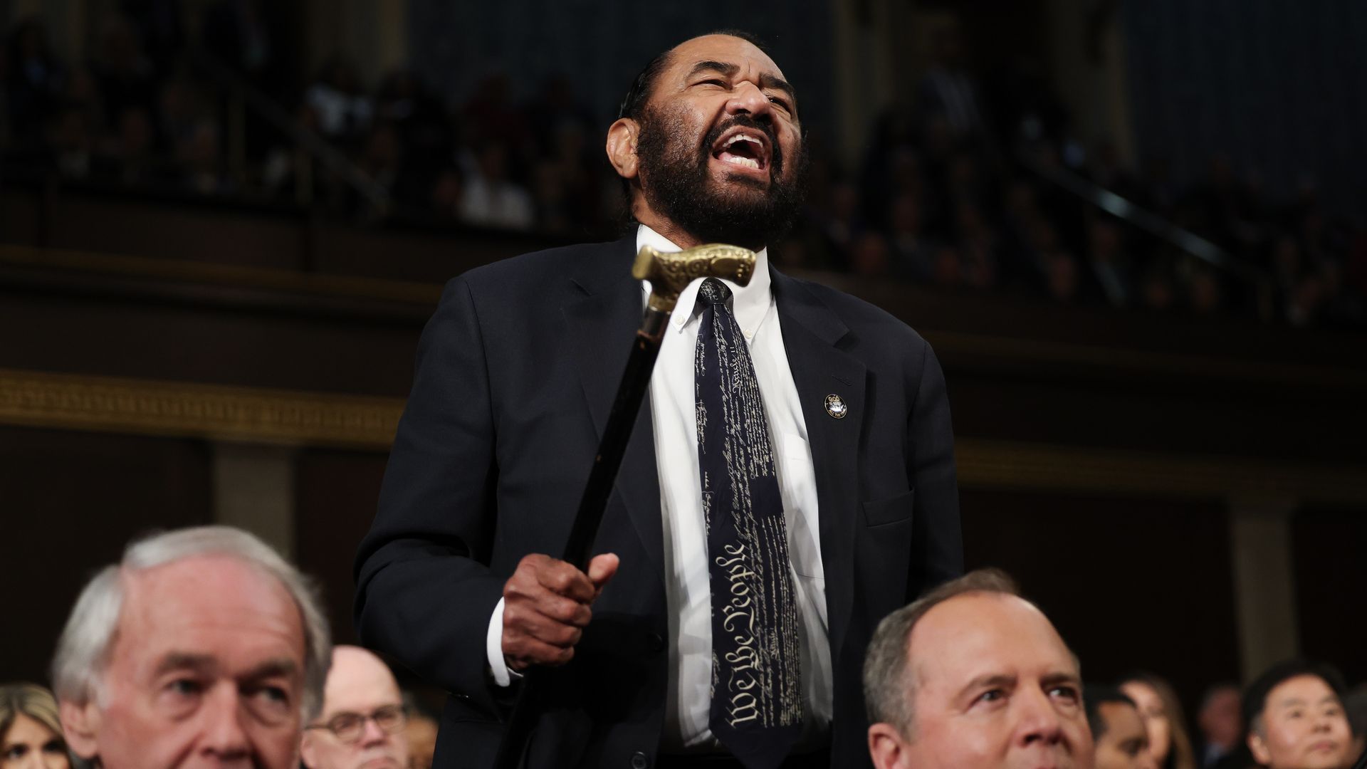 Rep. Al Green, wearing a gray suit and holding a gold-tipped cane, shouting while standing up in the filled House chamber.