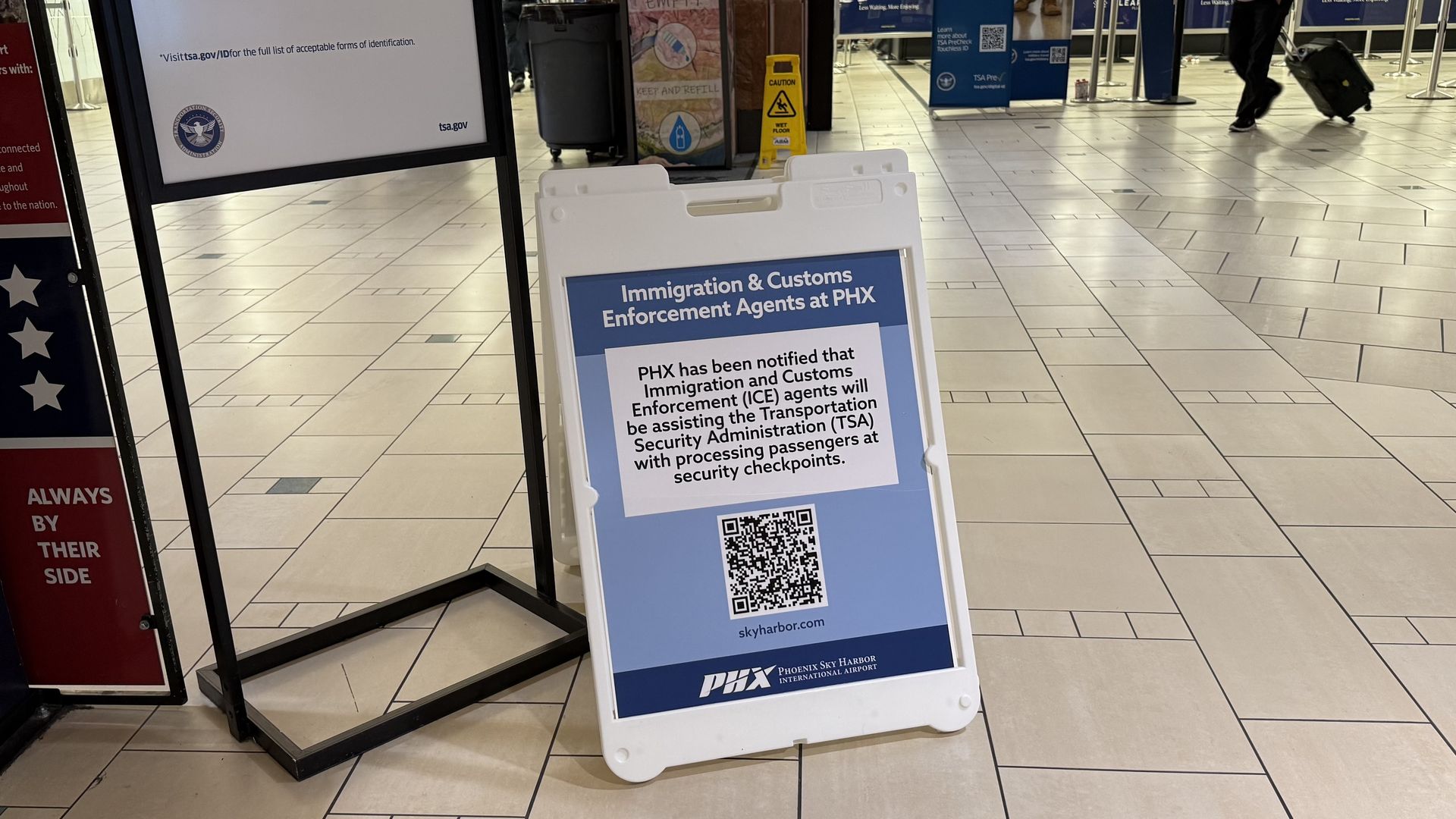 White A-frame sign at Phoenix airport reads "Immigration & Customs Enforcement Agents at PHX" with a QR code; a traveler with a wheeled suitcase walks across a tiled concourse.