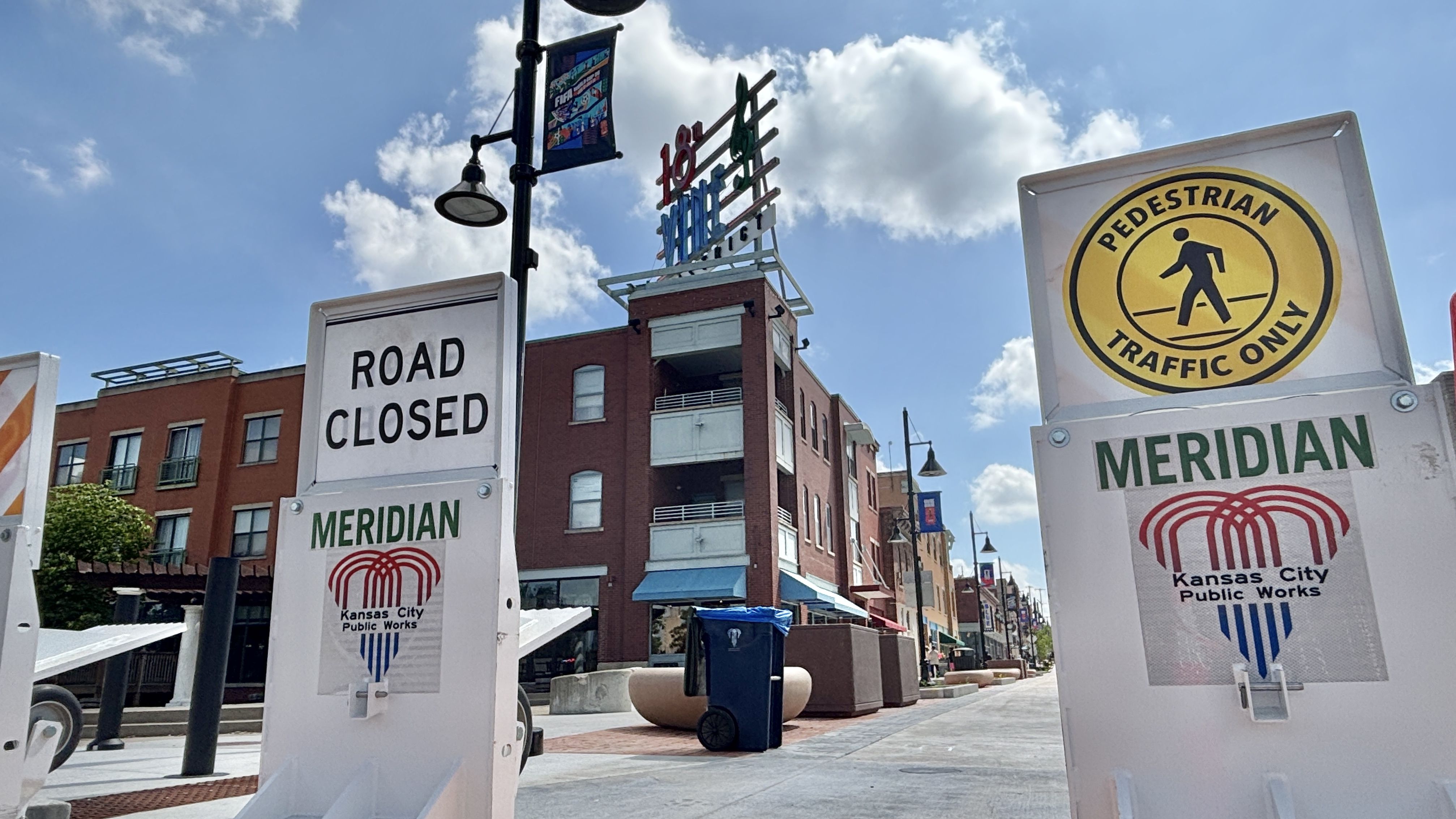Street scene with brick buildings under a blue sky. Foreground shows a large 'ROAD CLOSED' sign and a yellow 'PEDESTRIAN TRAFFIC ONLY' sign; banners and a building sign reading "18th and Vine" are visible.