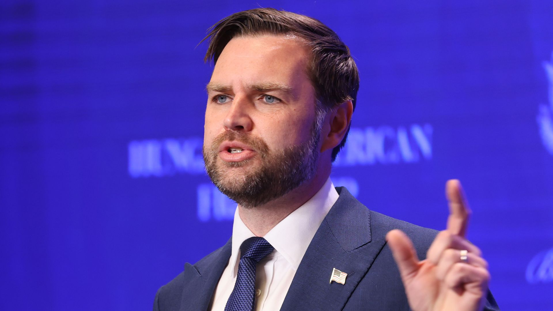 Vice President JD Vance, giving a speech with his left hand raised and pointing upward, and a US flag pin on his lapel. 