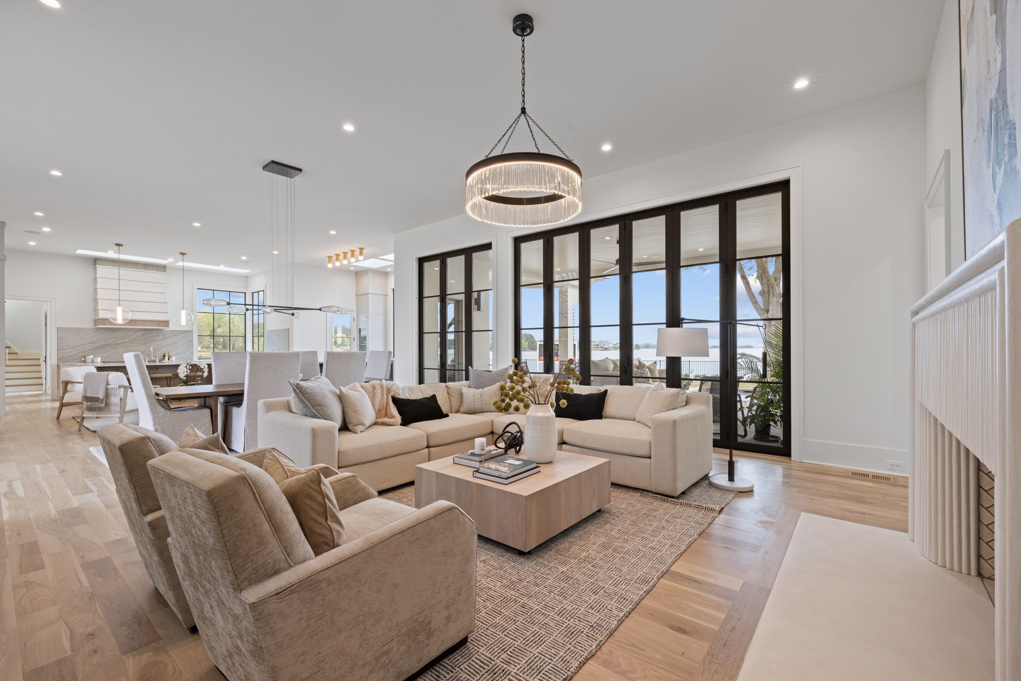 Bright modern living room with beige sectional sofa, two beige armchairs, wooden coffee table with vase, large glass doors with black frames, and open dining area with light wood floors.