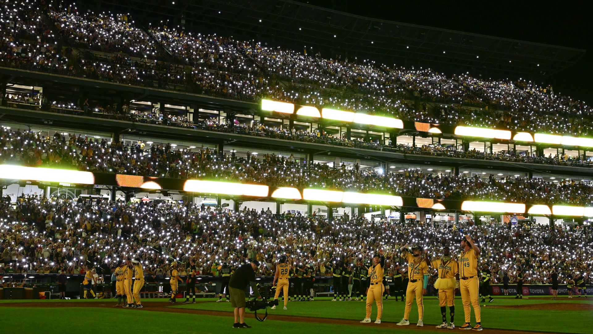 Night stadium crowded with fans in the stands, many holding up phone lights that glow white; on the baseball field, players in yellow uniforms stand on green grass.