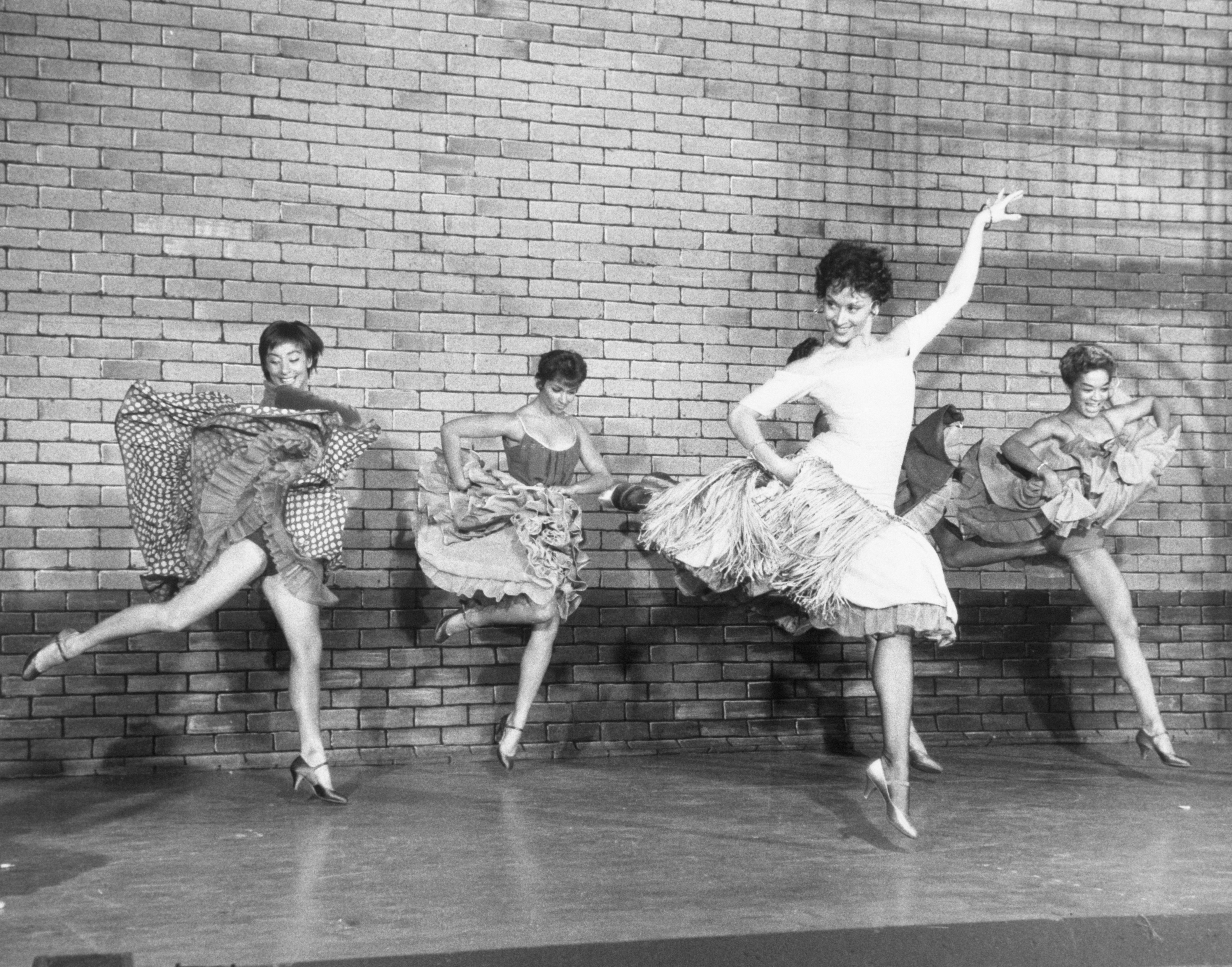 three dancers waving their skirts in the air dance behind Chita Rivera, who is holding one arm up while the other arm is at her waist. She is looking to the side and smiling. 