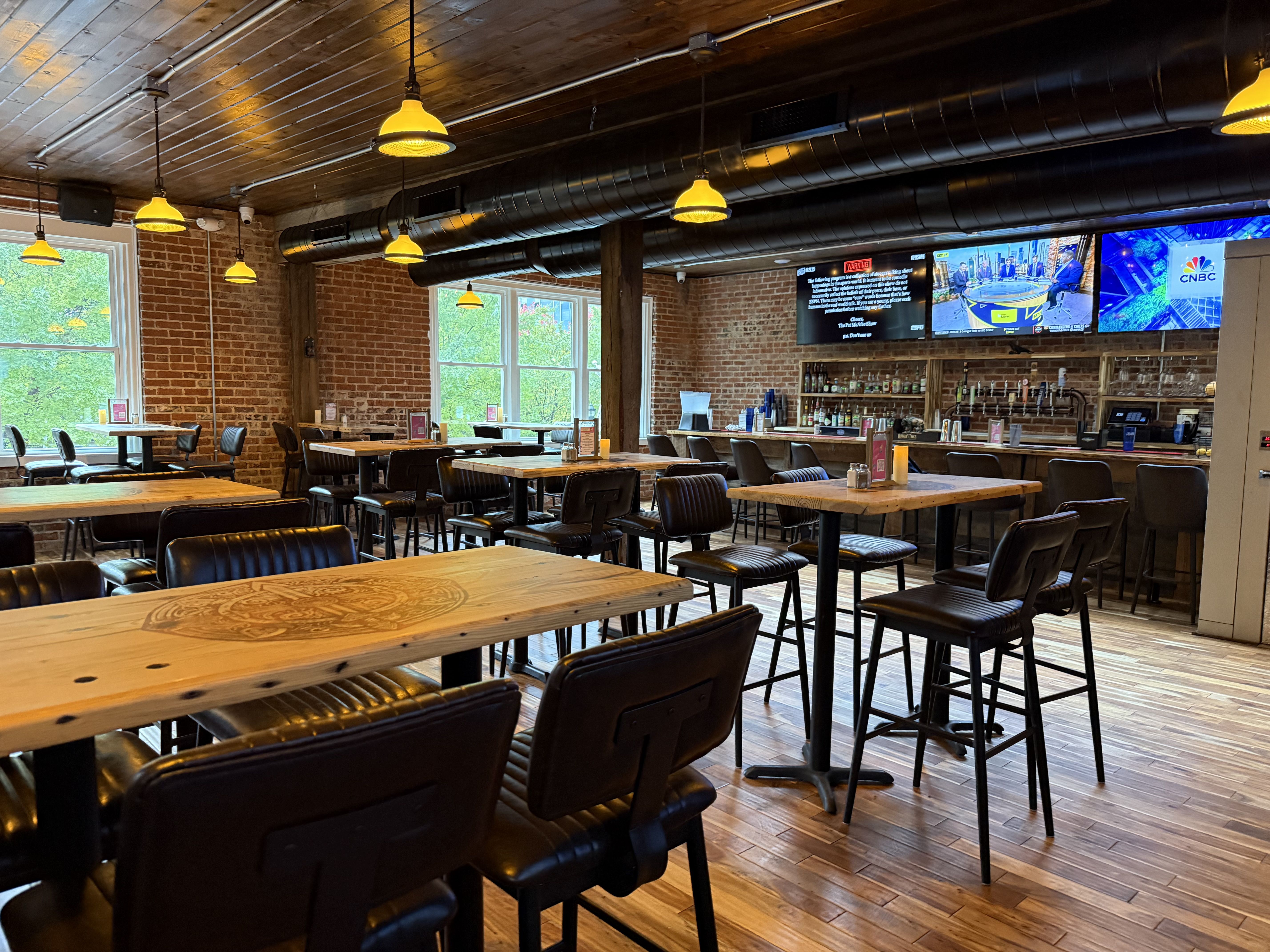 Empty bar with wooden tables and black chairs, brick walls, large windows with green trees outside, yellow hanging lights, and three TVs above the bar showing news and a logo.