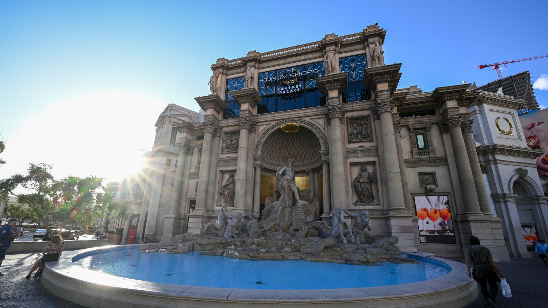 Sunlit, ornate building labeled "THE FORUM SHOPS" with statues and an arched entrance, and a circular fountain in the foreground; blue sky and a construction crane to the right.