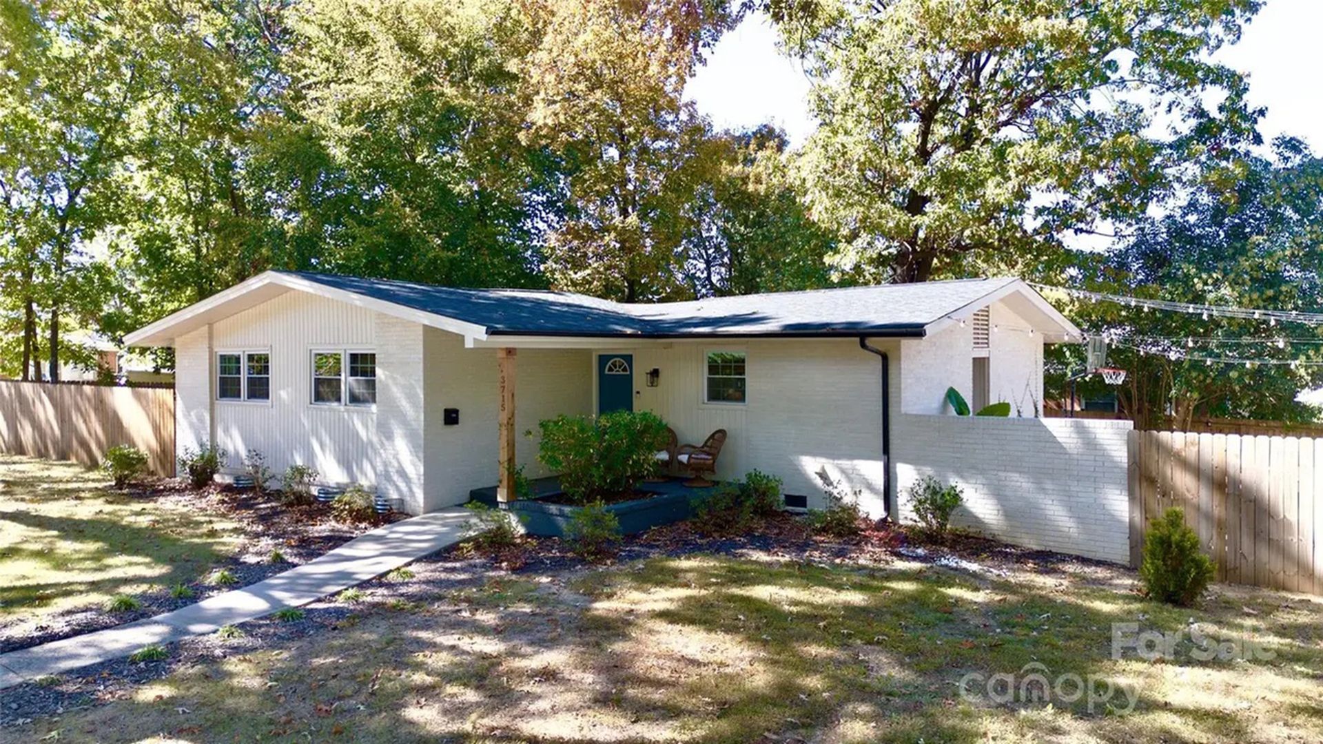 Single-story white brick house with a blue door, two chairs on the porch, surrounded by trees and a fenced yard on a sunny day.