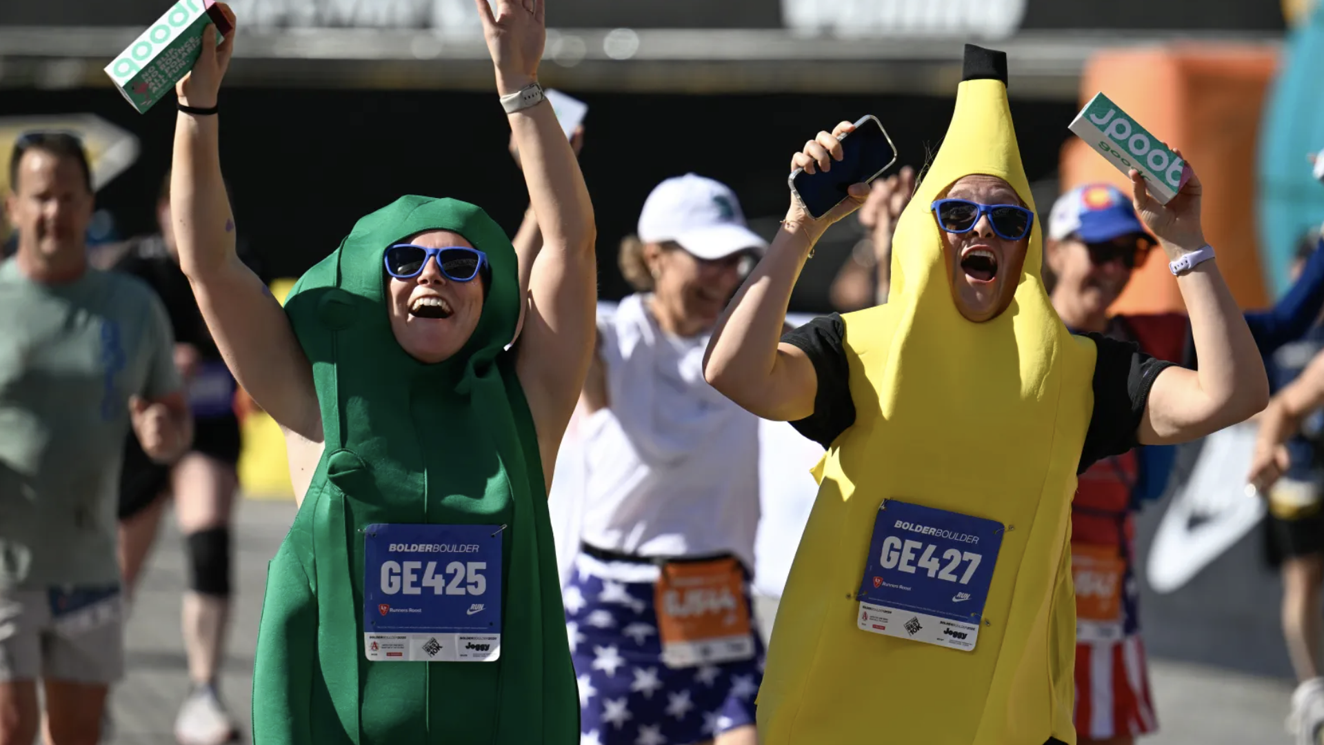 Costumed runners cheer at a race: a green leafy suit and a yellow banana suit, both with blue sunglasses. They raise arms and hold drink boxes; bibs read GE425 and GE427.