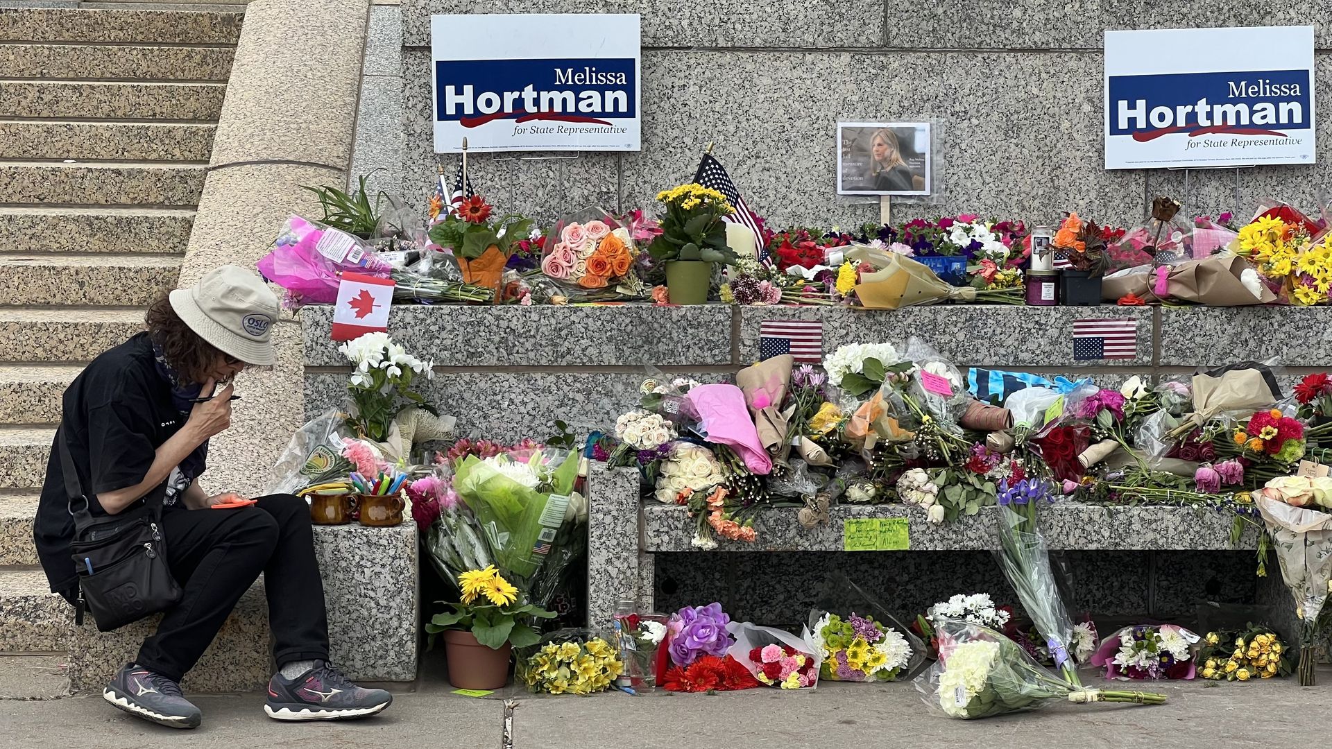 Person wearing a beige hat sits on granite steps beside a memorial of bouquets and signs reading "Melissa Hortman for State Representative," with small American flags among the flowers.
