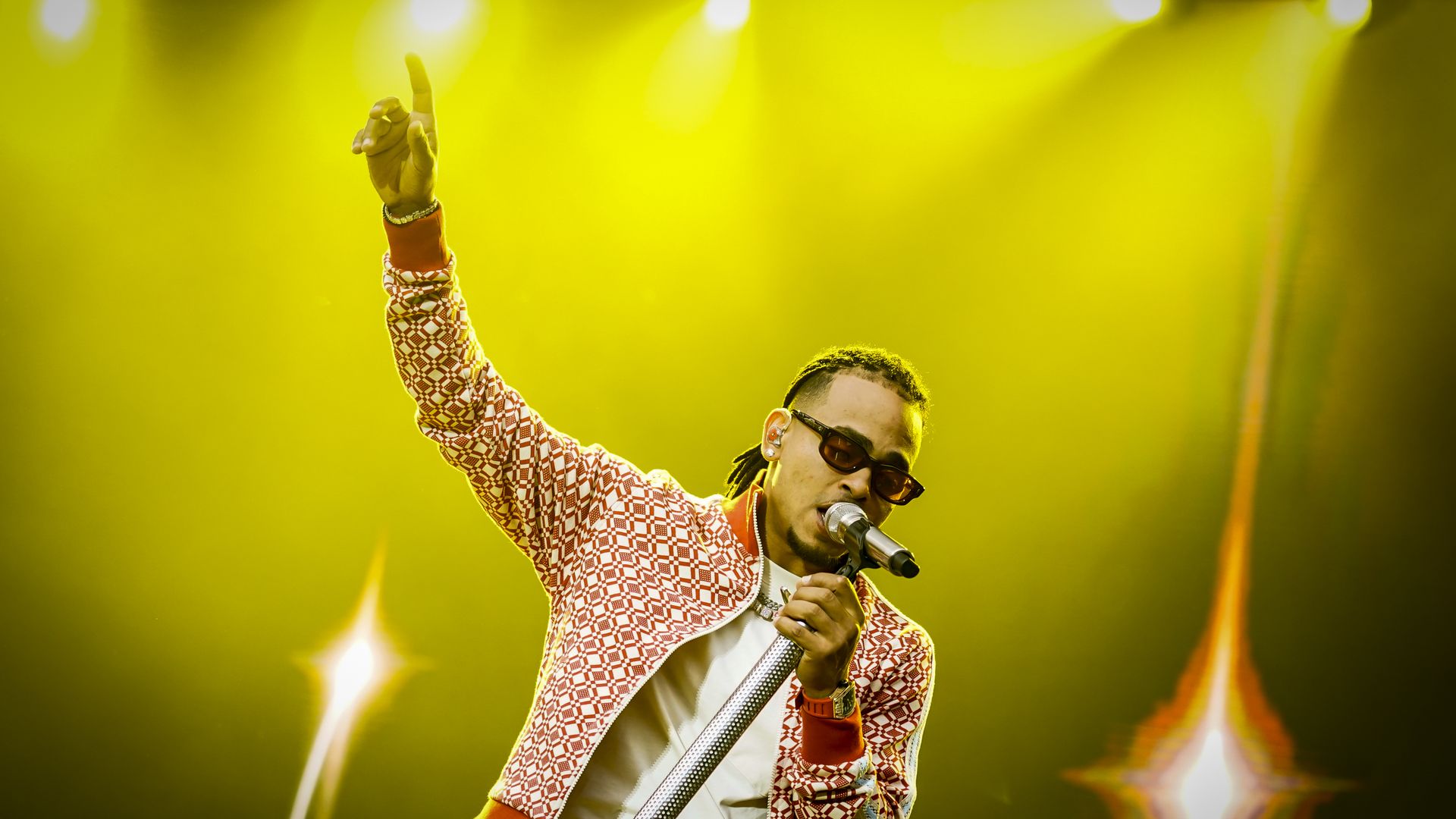 Ozuna wears a red-and-white patterned jacket and sunglasses as he points to the sky. 